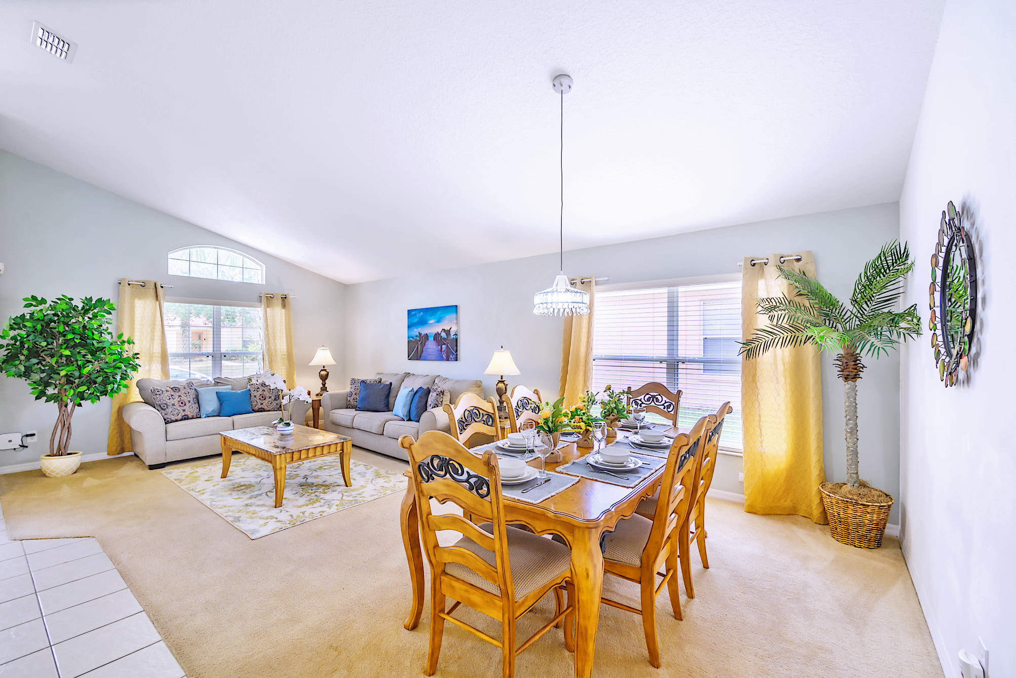 Open-concept living and dining area with light carpet, featuring a beige sectional sofa, a wooden coffee table, and a dining table set for six with wooden chairs.