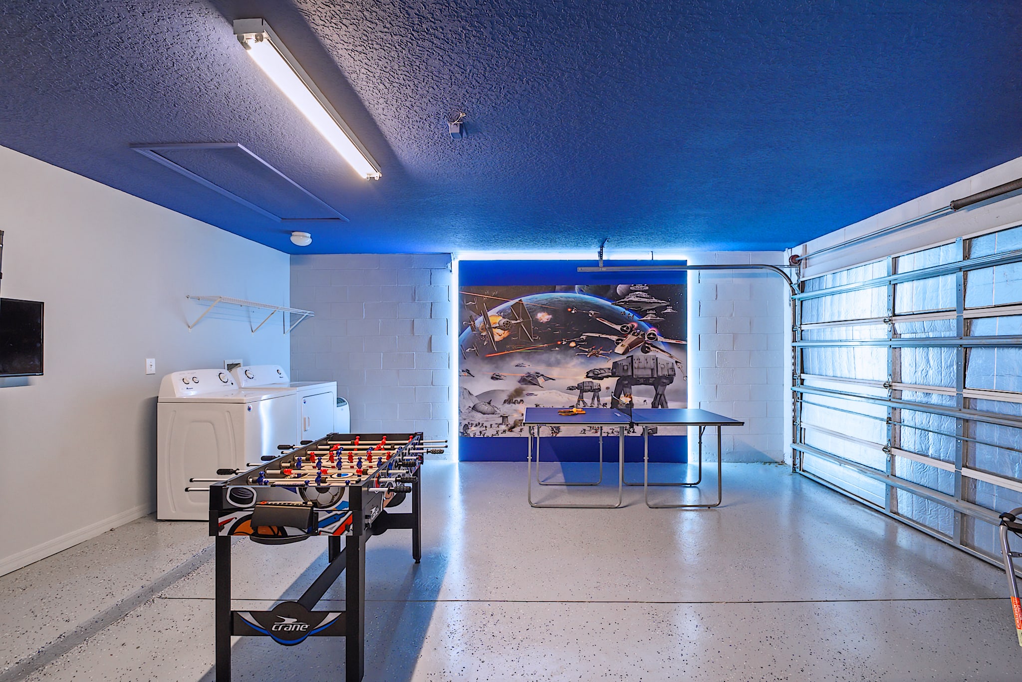 Game room with a blue ceiling and a large Star Wars mural on the wall, featuring a foosball table and a small table, with a washer and dryer visible in the background.