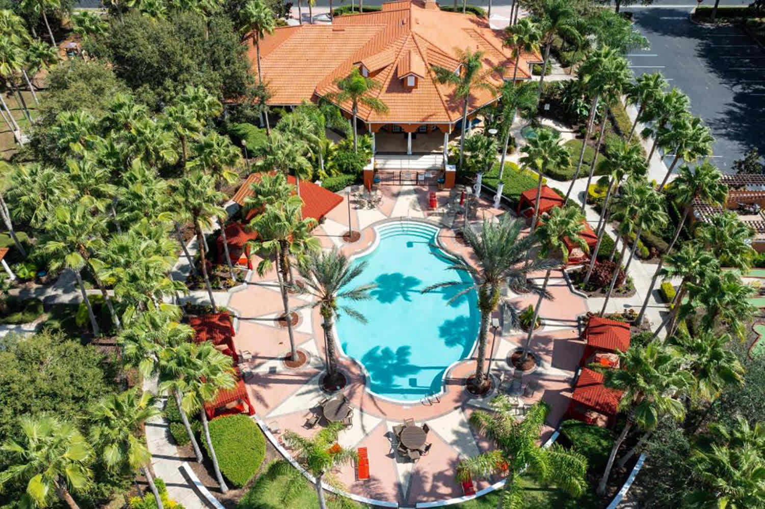 Aerial view of a large communal pool area surrounded by numerous palm trees and lounge chairs, with a central building featuring a red tile roof.