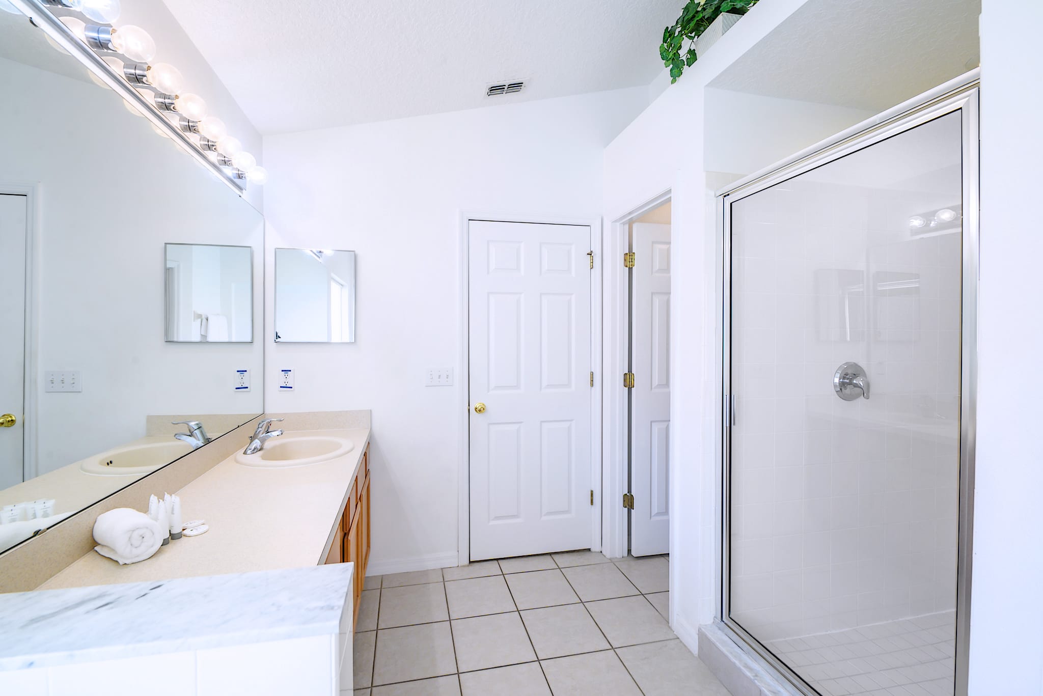 Bright, spacious bathroom featuring a double vanity with light countertops, two sinks, and a large mirror, alongside a glass-enclosed walk-in shower with white tiled walls.