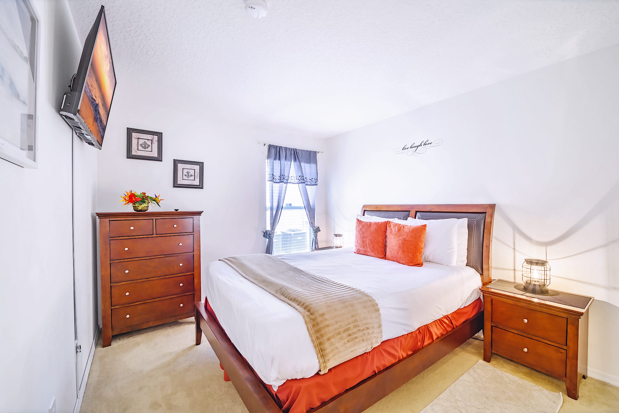 Cozy bedroom with a dark wood bed frame, white and orange bedding, a matching dresser, and a wall-mounted TV, all set against white walls with a decorative script above the bed.