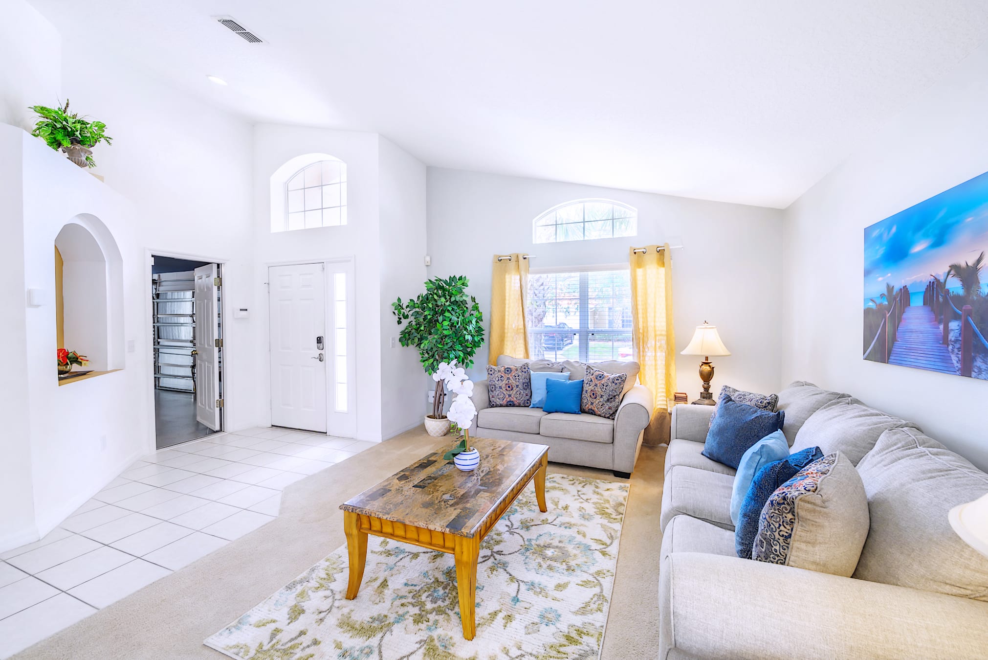 Bright living room with white walls, vaulted ceilings, and arched windows, featuring two light-colored sofas with blue accent pillows, a wooden coffee table on a floral rug, and a large wall art depicting a beach scene.
