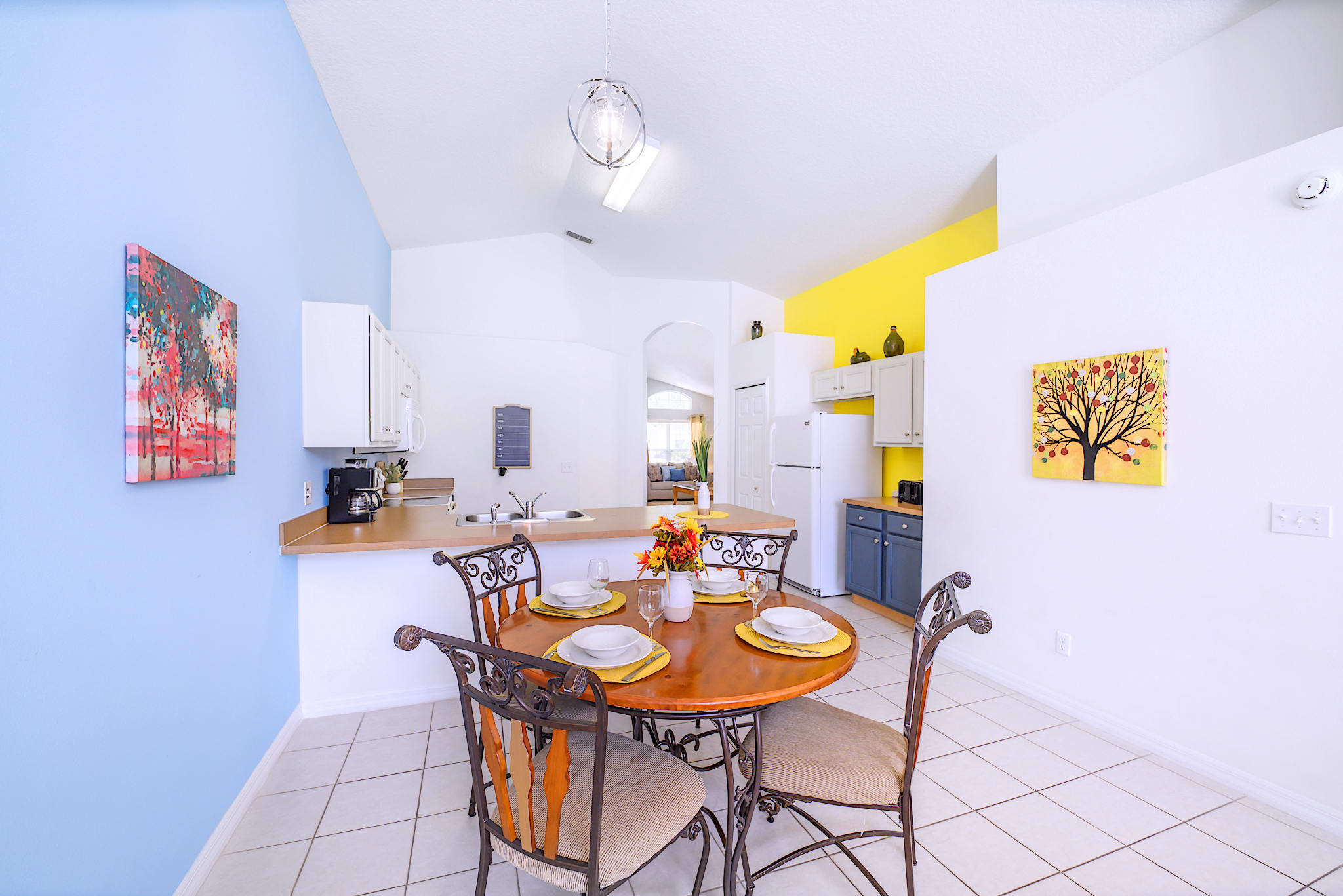 Casual breakfast nook with a round wooden table and four chairs, situated between light blue and yellow accent walls, offering an open view into the kitchen area.