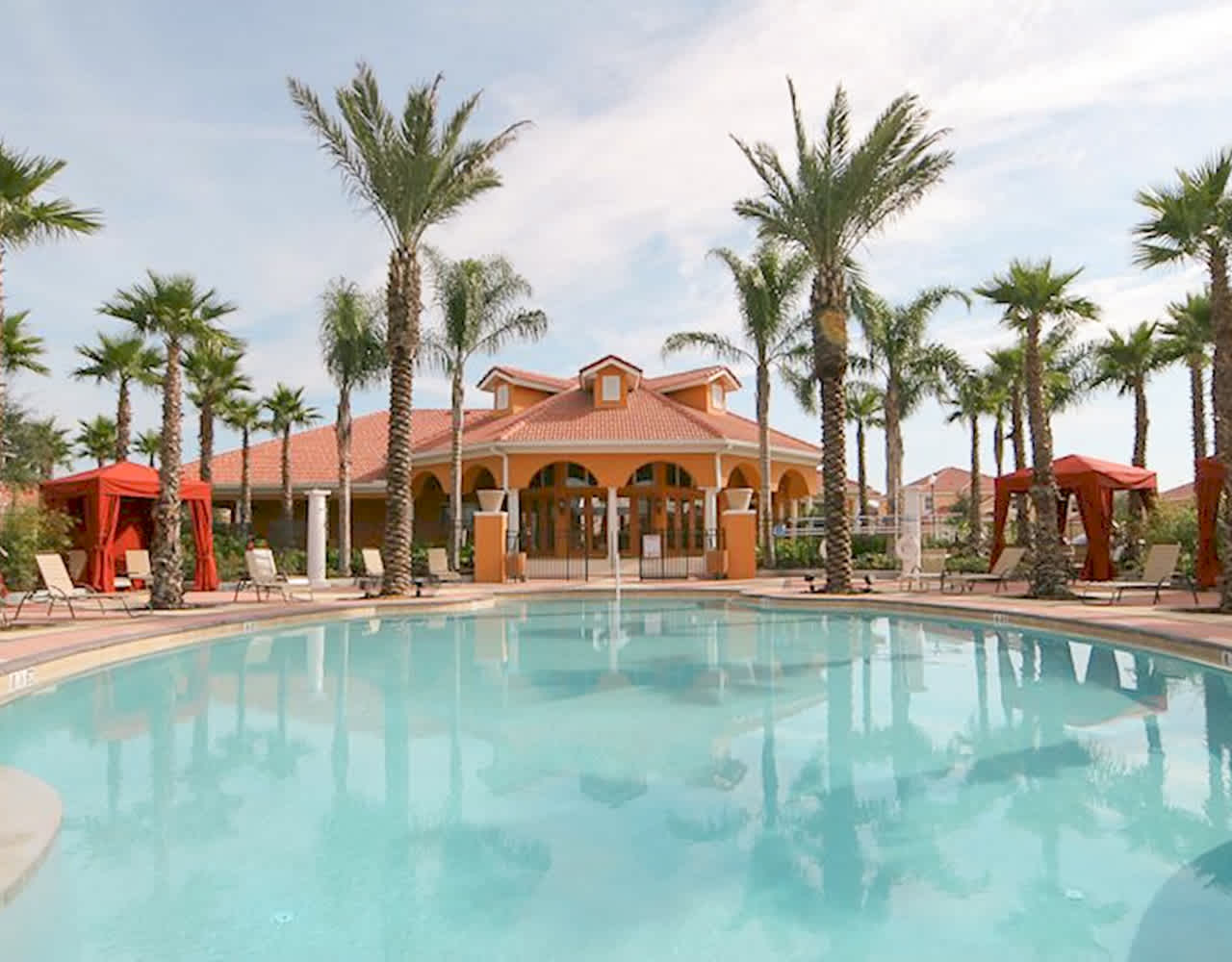 Expansive communal outdoor swimming pool surrounded by numerous tall palm trees, with a large clubhouse building featuring an orange roof in the background under a clear sky.
