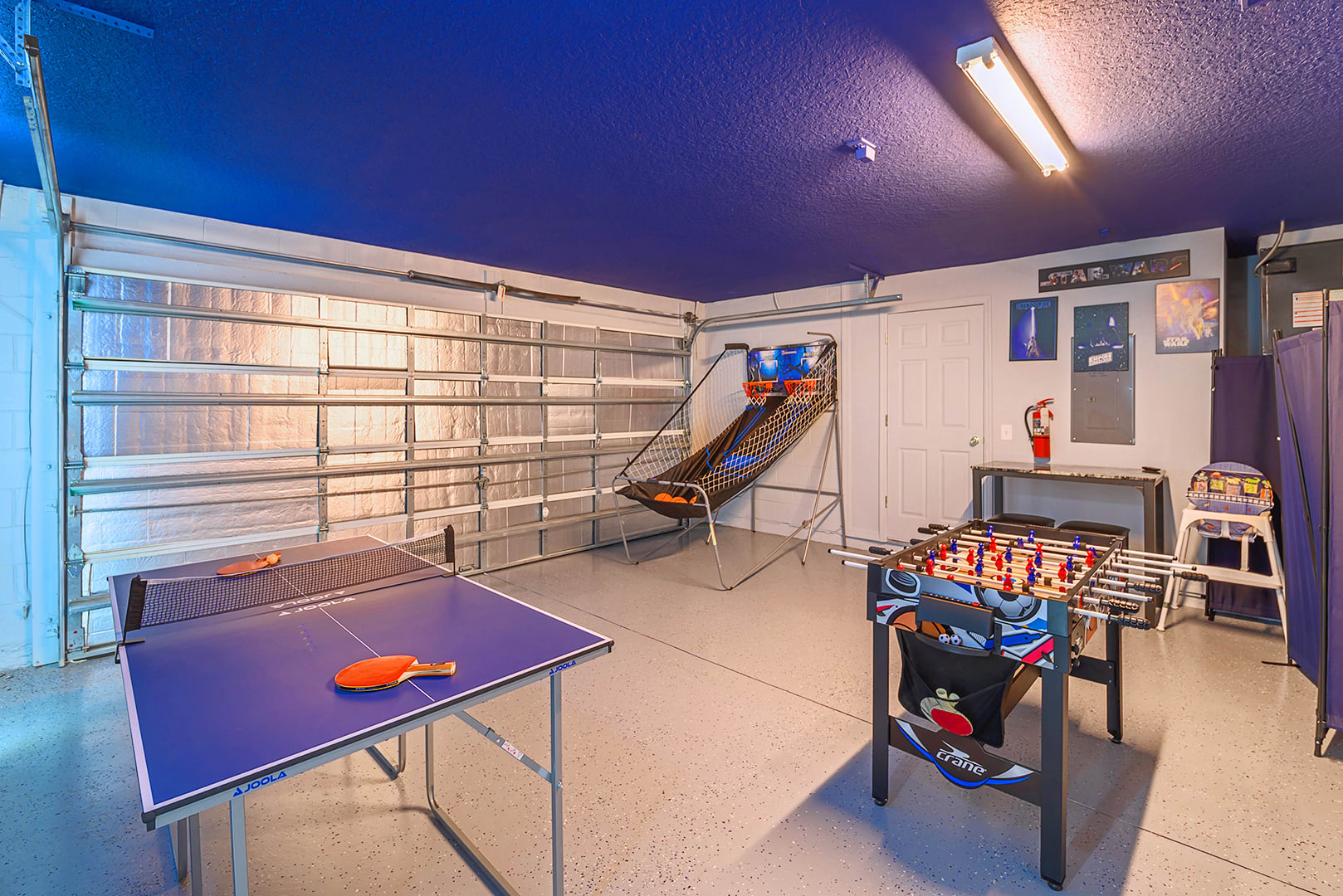 Garage converted into a game room with a blue ceiling, featuring a ping-pong table, a foosball table, and a basketball arcade game, with Star Wars posters on the wall.
