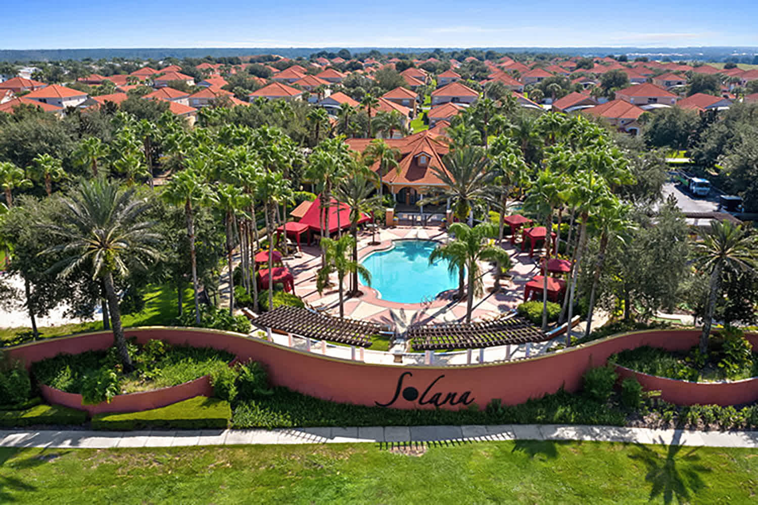 Aerial view showcasing the grand entrance to the 'Solana' resort community, featuring a large decorative sign, manicured green lawns, and numerous palm trees. In the background, a communal pool area and a vast residential area with red-roofed homes are visible under a clear blue sky.