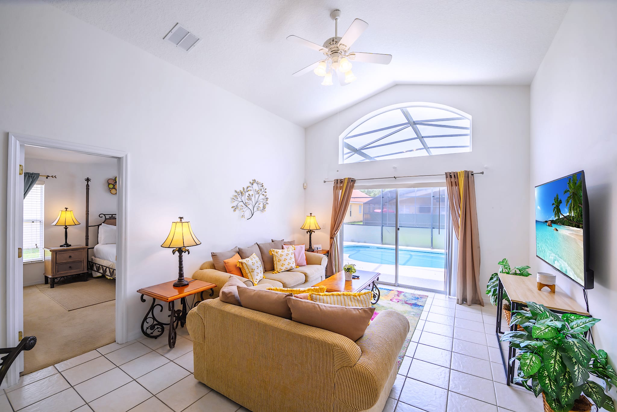 Cozy living room with light-colored sofas, a wooden coffee table, and a large flat-screen TV, featuring a vaulted ceiling with a fan and a wide arched window/sliding door overlooking a private pool area.