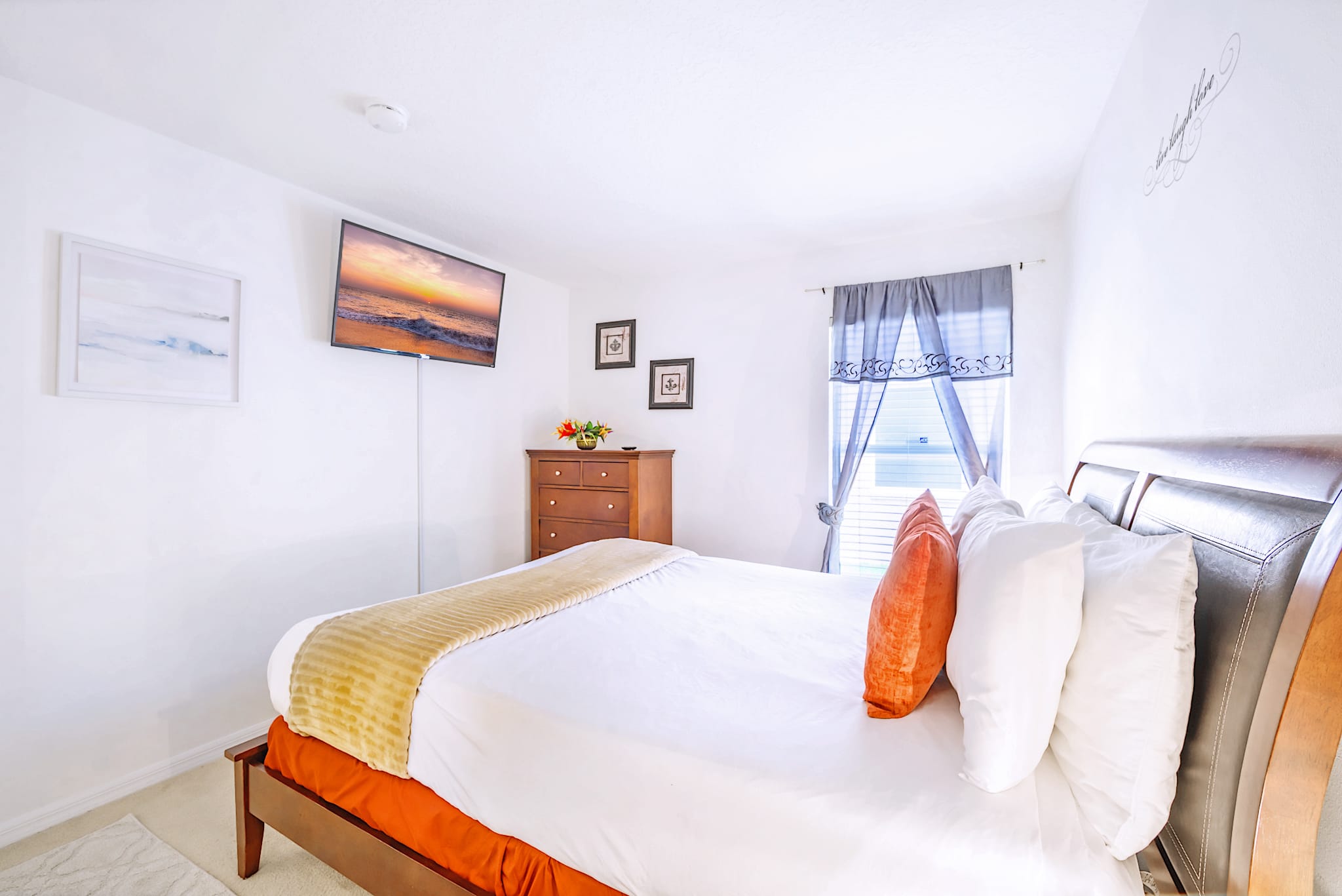 View of a comfortable bedroom featuring a large bed with white and orange bedding, a dark wood nightstand with a lamp, and a wall-mounted TV, with a window dressed in dark blue curtains.