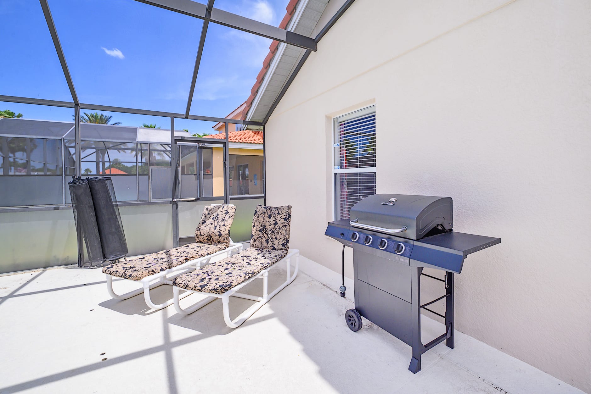 Close-up of a black BBQ grill and two white lounge chairs with patterned cushions on a light concrete patio, enclosed by a screen.