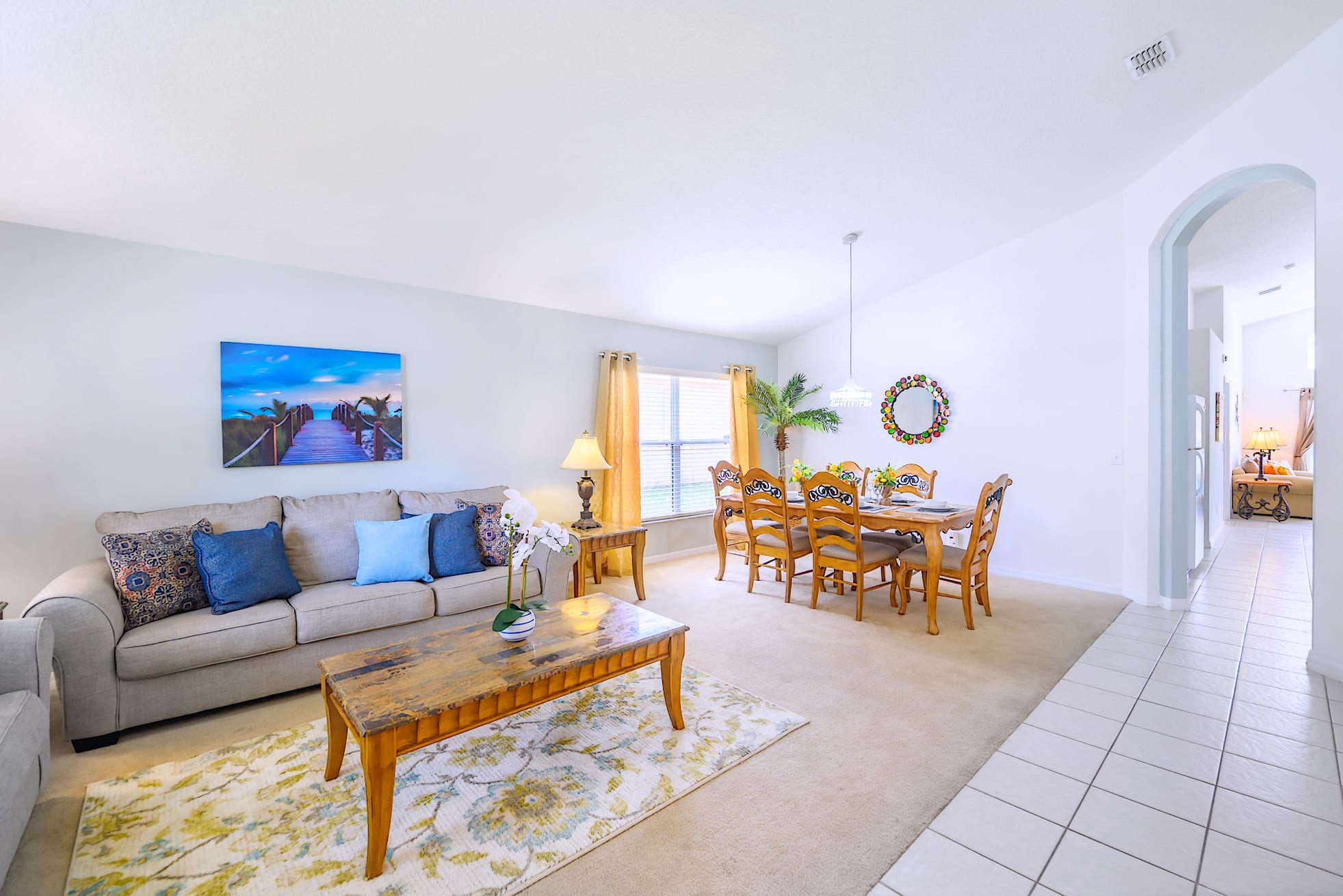 Expansive living and dining area with light-colored walls, featuring a beige sofa with blue accent pillows, a wooden coffee table on a floral rug, and a dining table with six chairs under a chandelier, next to a window with yellow curtains.