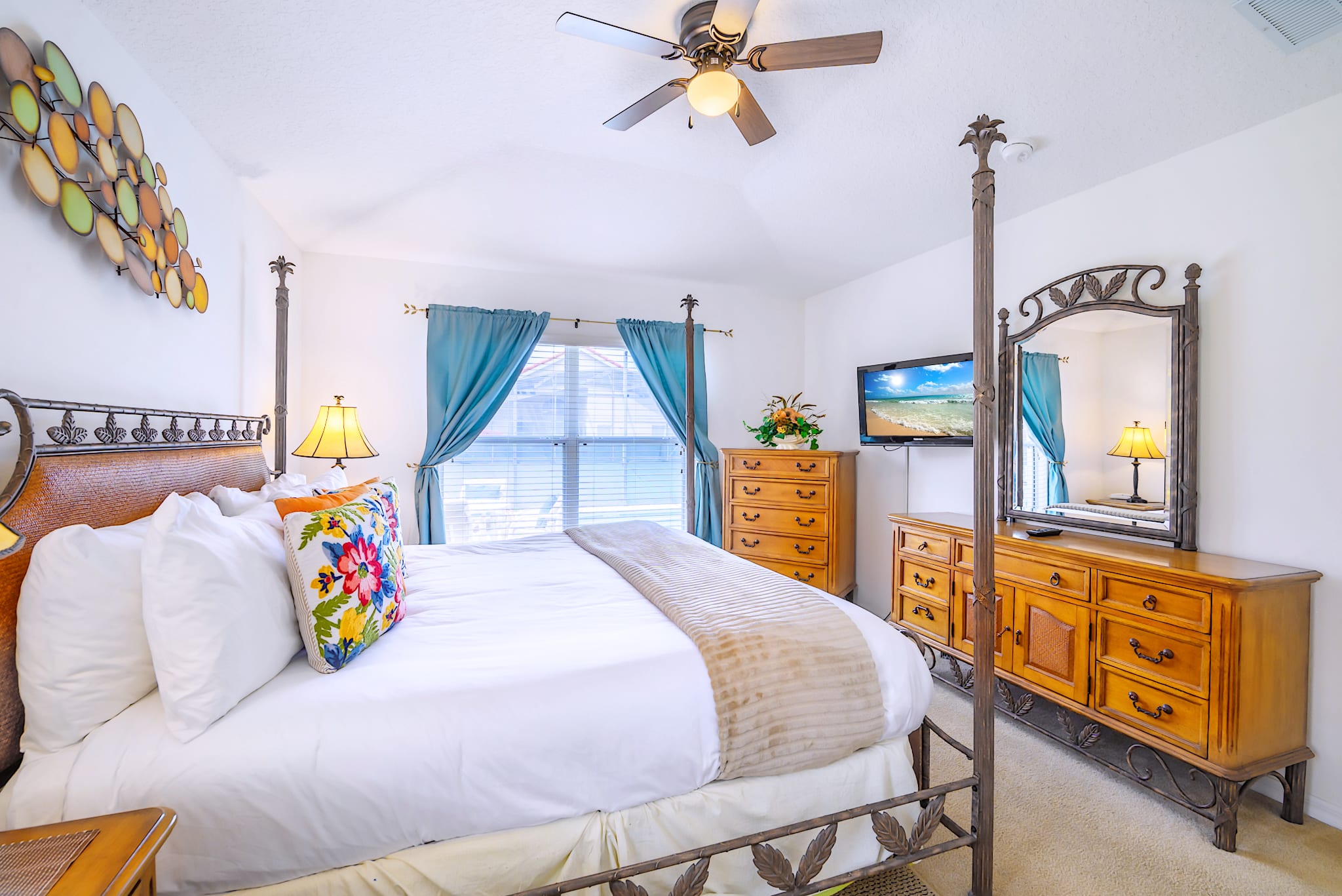 Spacious bedroom featuring a large four-poster bed with a wicker headboard, wooden dressers, a wall-mounted TV, and a ceiling fan, with a window dressed in blue curtains.