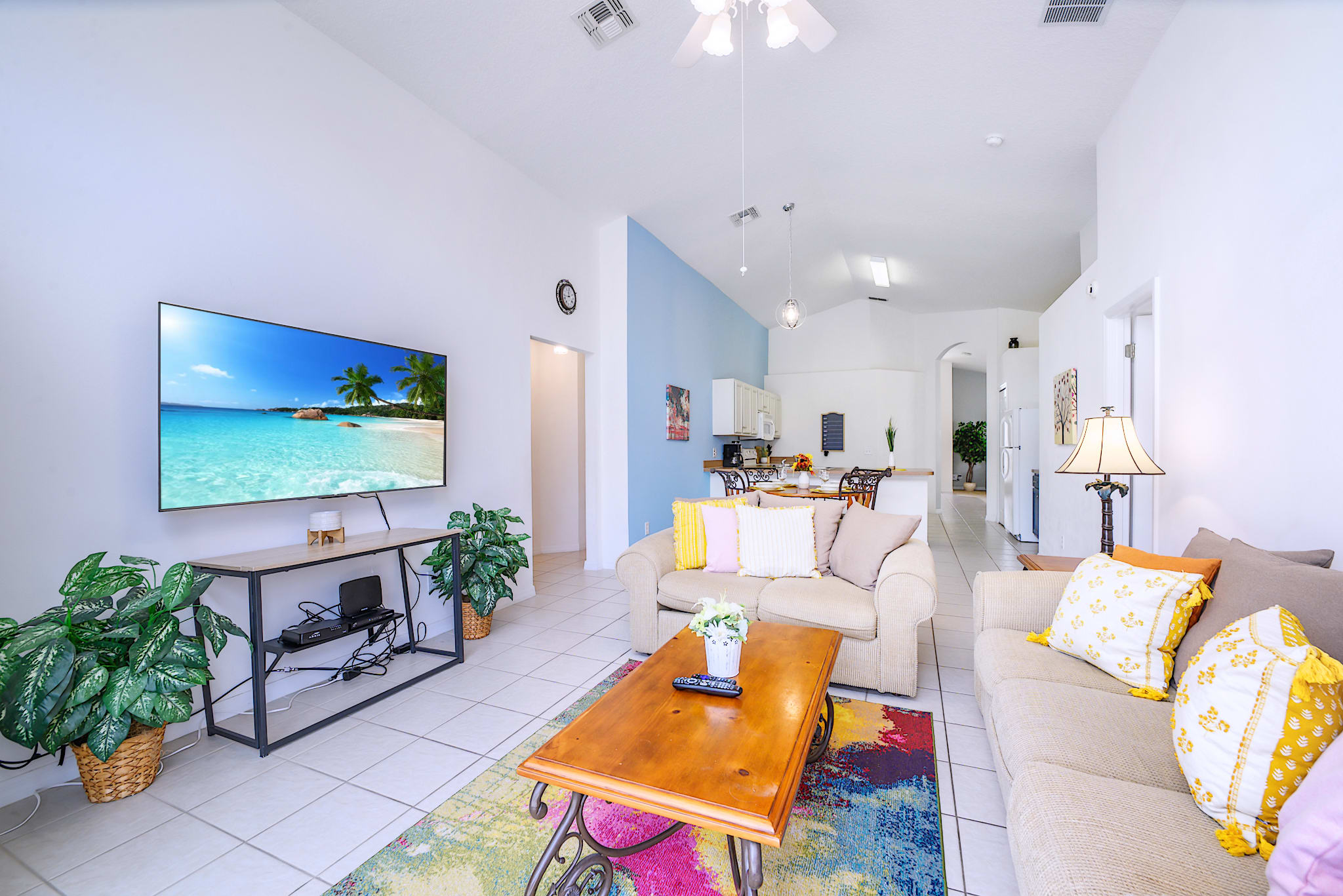 Bright living room with two light-colored sofas adorned with colorful pillows, a wooden coffee table, and a large wall-mounted TV, connected to an open-concept kitchen with a light blue accent wall.