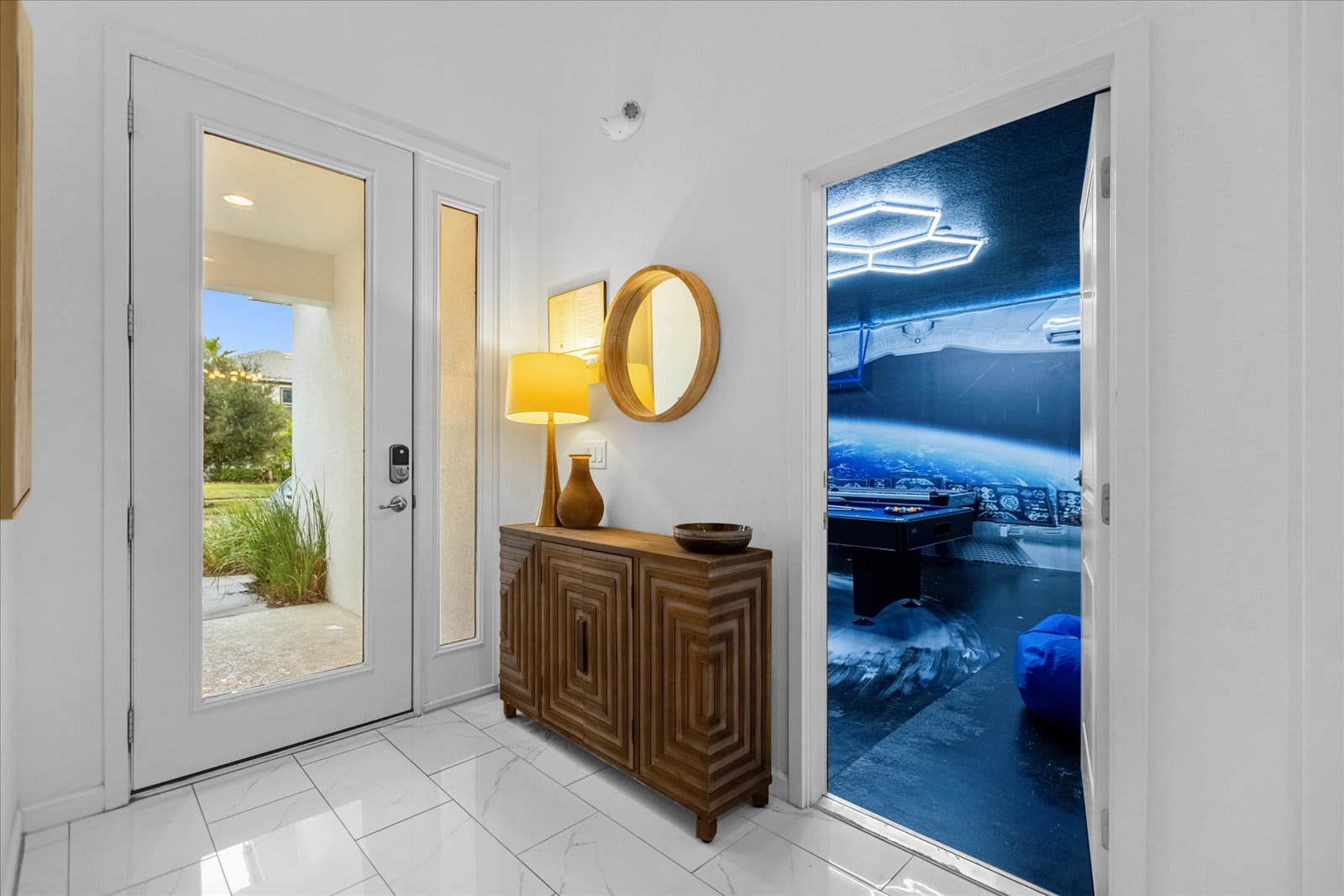 Bright entryway with white tile flooring and walls, featuring a wooden console table with a lamp and a round mirror. A glimpse into the Star Wars-themed game room is visible through an open doorway, hinting at the fun within.