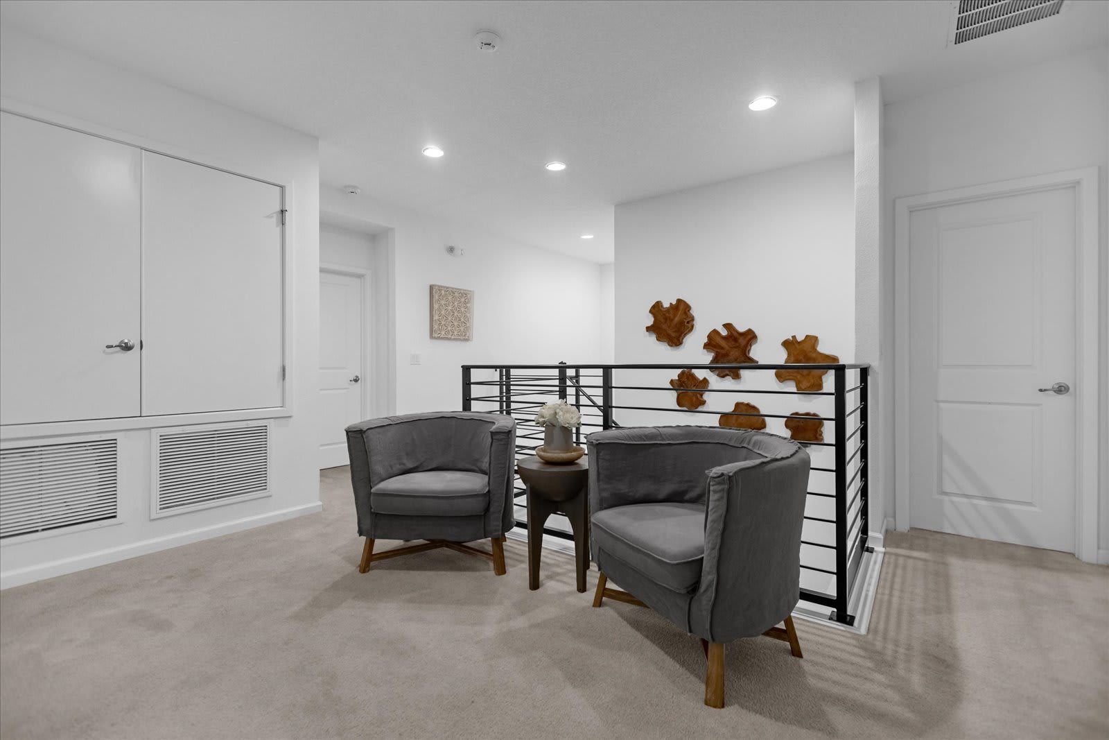 Upper landing area with light gray carpet, featuring two comfortable gray armchairs and a small side table, overlooking a staircase with black railings, and decorated with abstract wooden wall art.