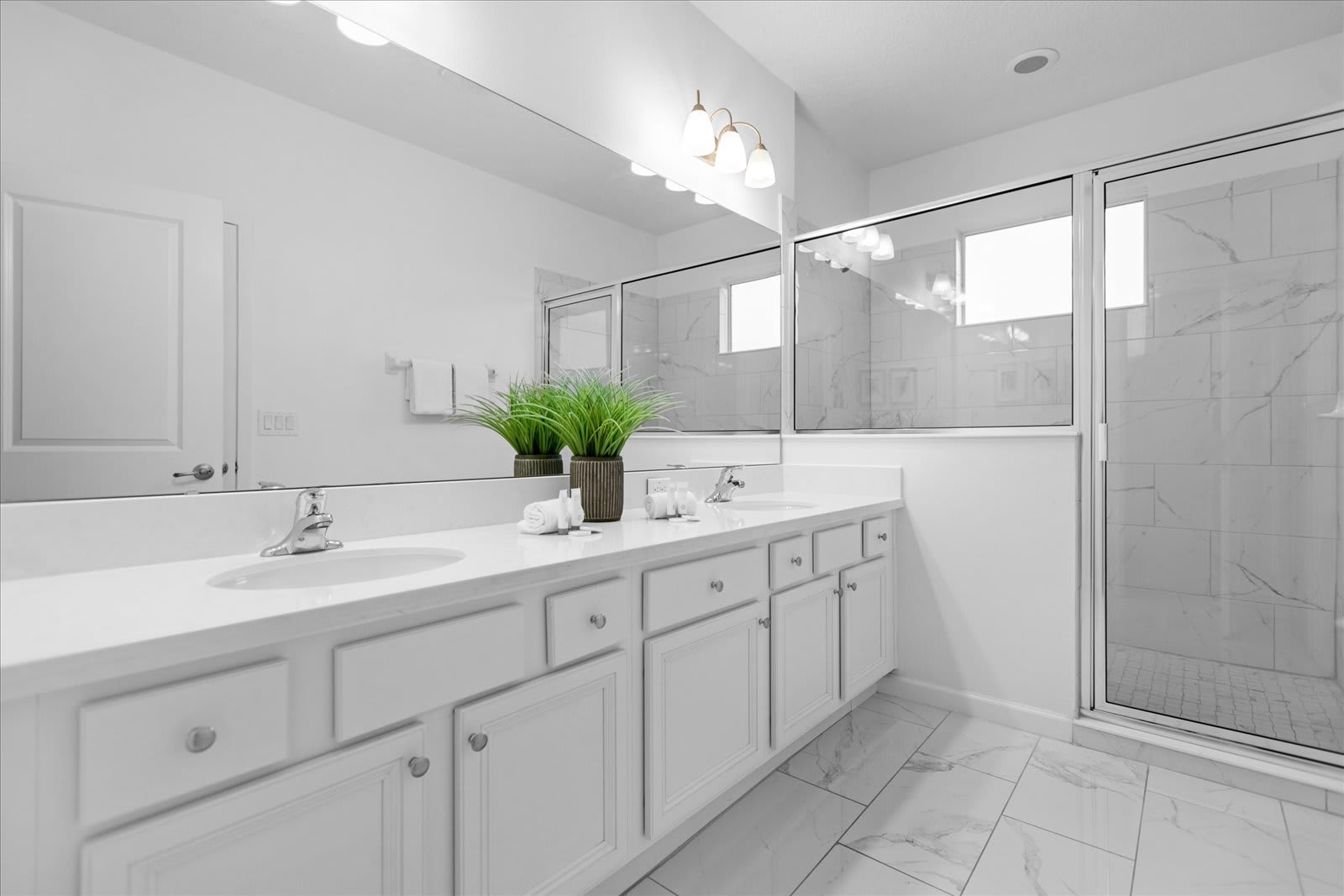 Modern white bathroom with a long double vanity, white countertops, white cabinets, and a large mirror. A glass-enclosed shower is visible on the right, and a separate toilet room is reflected in the mirror.