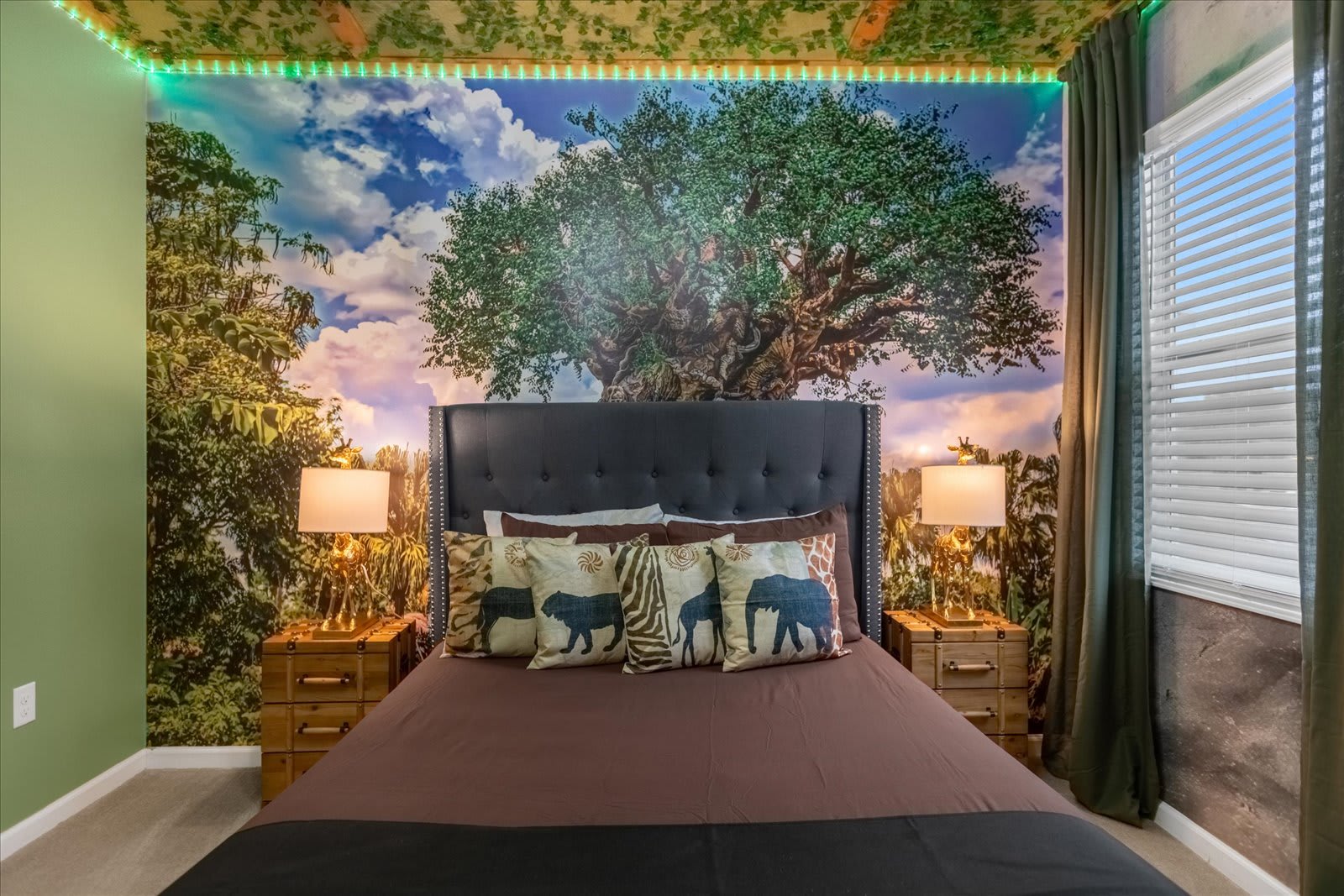 Themed bedroom with a large bed featuring a dark tufted headboard and animal print pillows, set against a wall mural of a large tree and sky, with a jungle-themed ceiling and green accent lighting.
