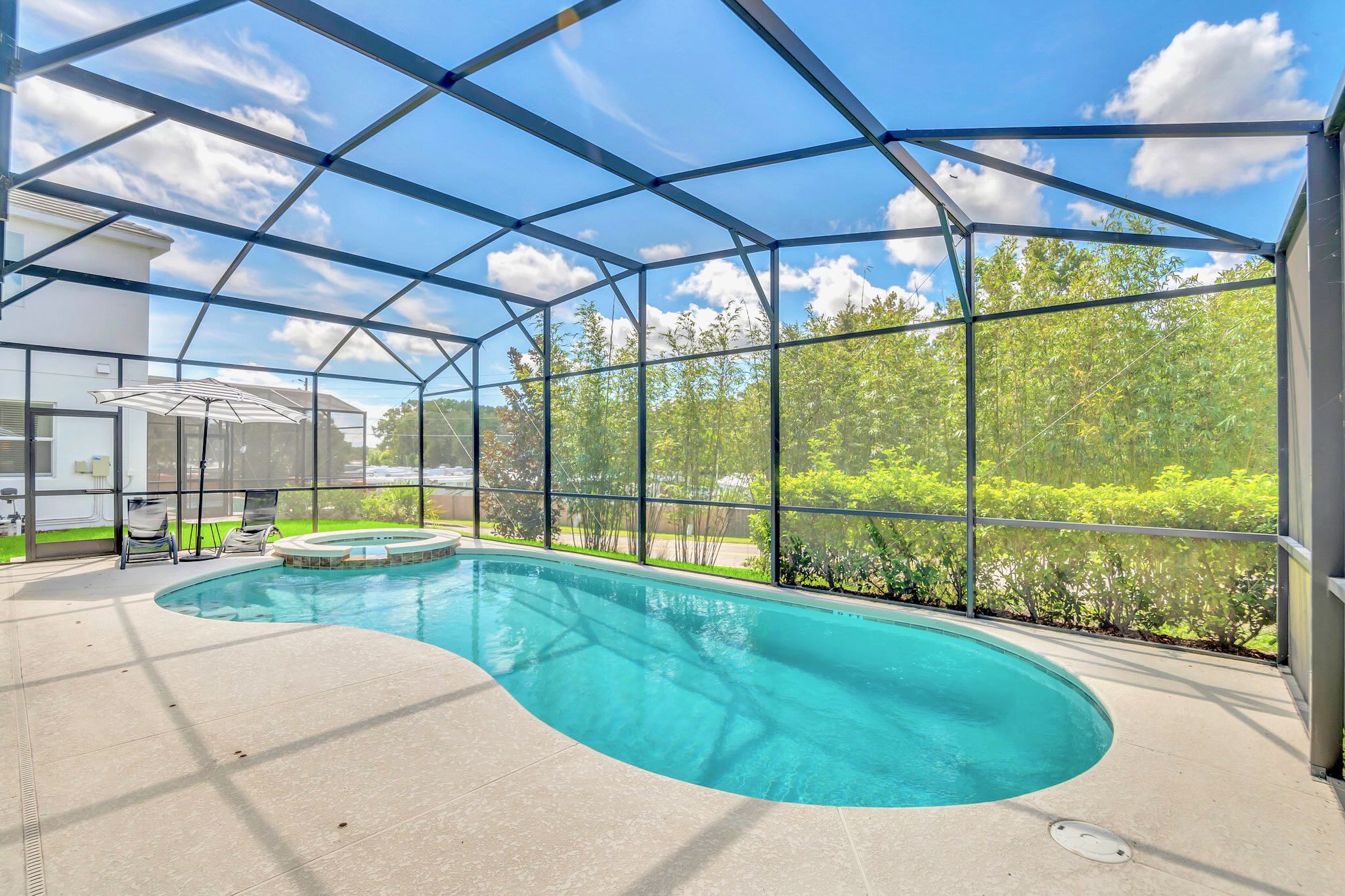 Expansive screened-in private pool area with a freeform pool and integrated hot tub, surrounded by light-colored patio decking. Tall green foliage provides privacy beyond the screen enclosure under a bright blue sky.