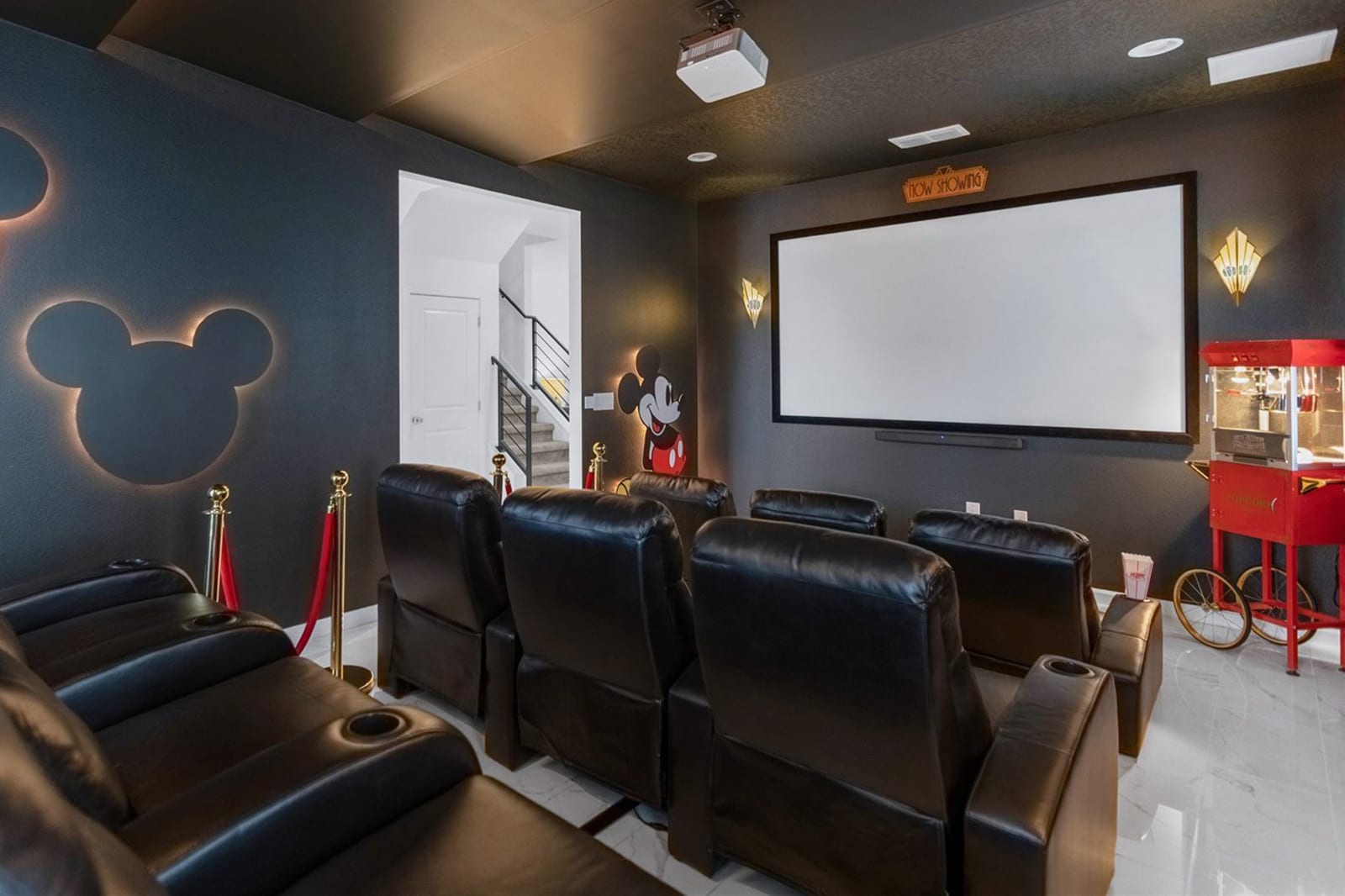 A dark, luxurious home cinema room featuring multiple rows of black leather recliners, a large projection screen, a vintage red popcorn machine, and subtle Mickey Mouse-themed wall lighting.