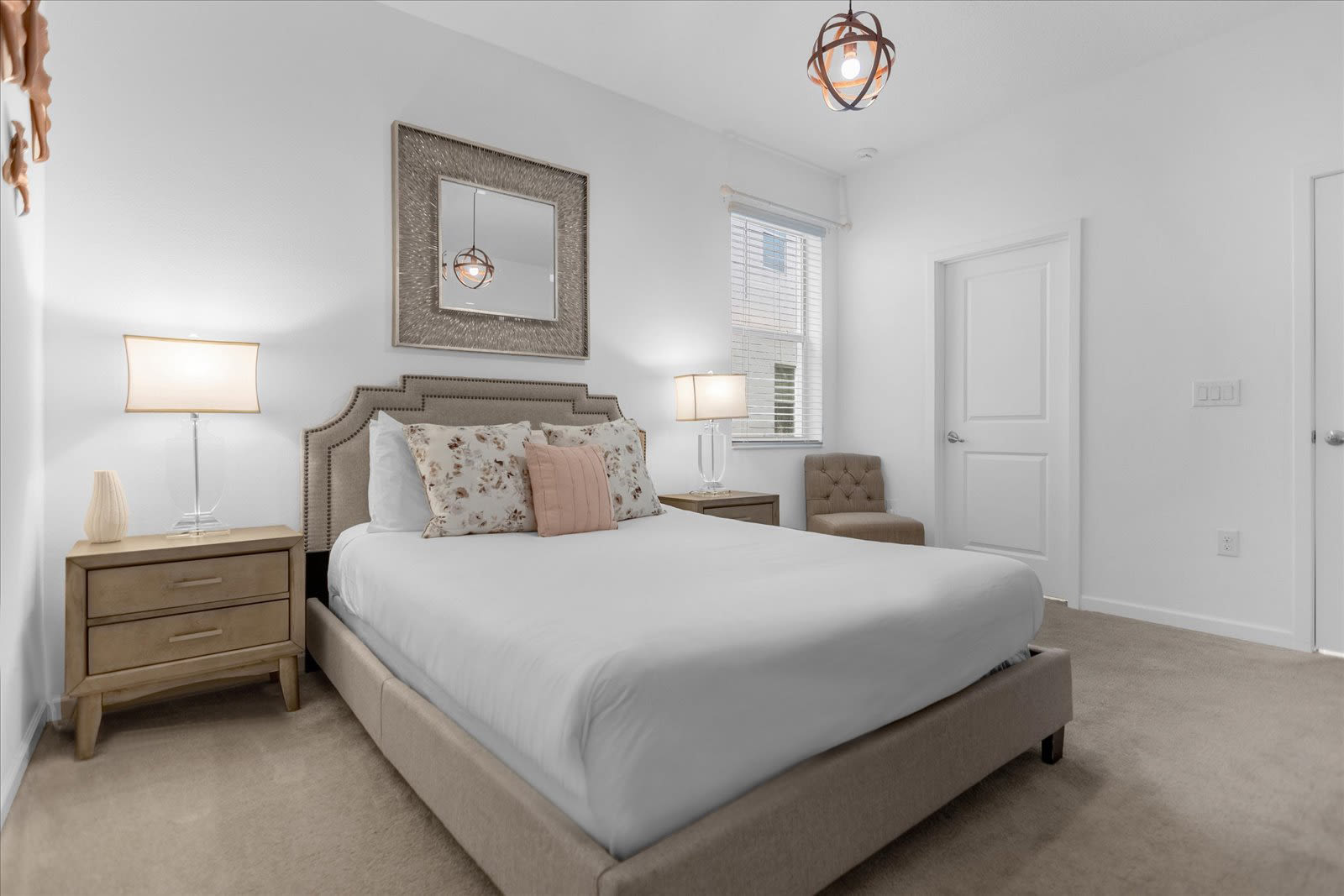Bright bedroom with a queen-sized bed, a tufted headboard, two nightstands with lamps, and a large decorative mirror above the bed. The room has carpeted flooring, a window with blinds, and a closet with bi-fold doors.