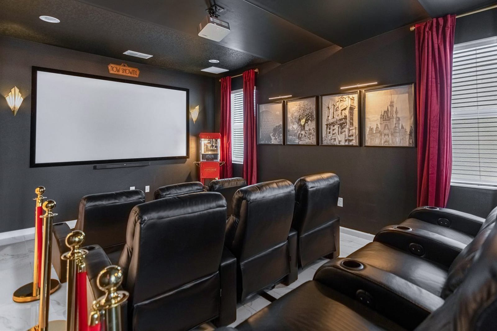 Dark-walled home cinema with multiple rows of plush black leather recliners, a large projection screen, and a vintage red popcorn machine. Gold stanchions and red velvet ropes add a classic movie theater ambiance.