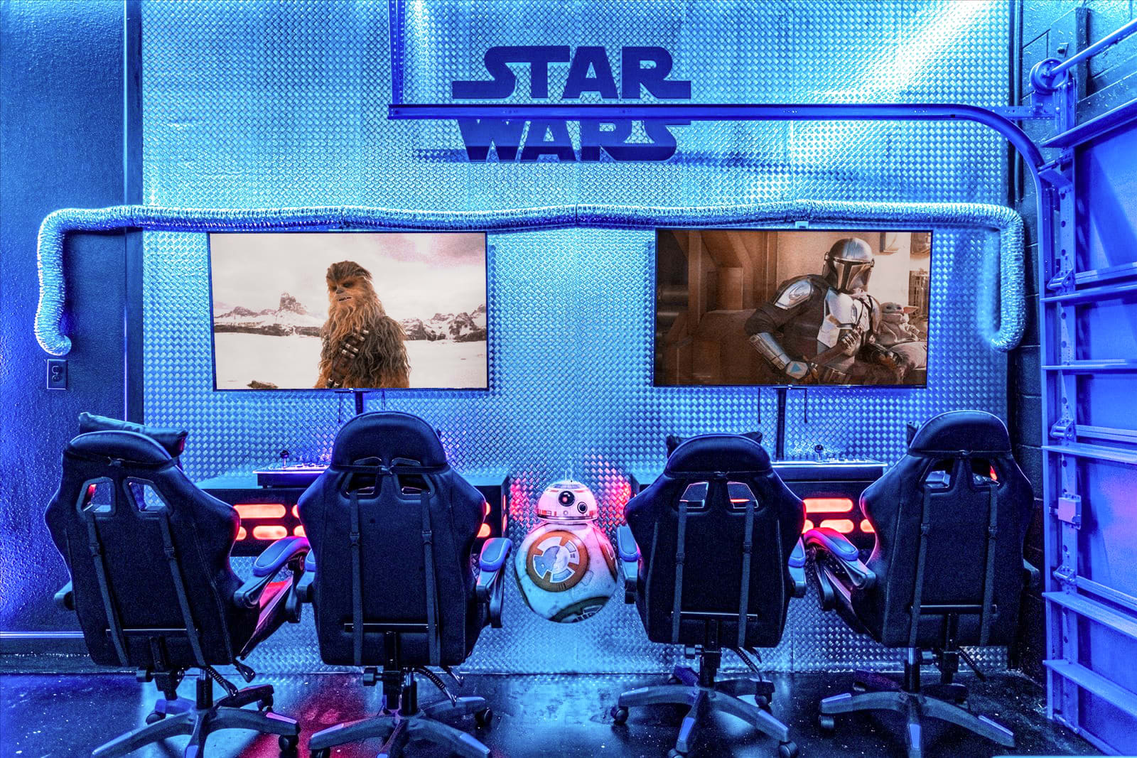 Close-up of the Star Wars-themed gaming area with two large screens displaying characters, four black gaming chairs, and a BB-8 droid figure, all set against a blue-lit metallic wall.