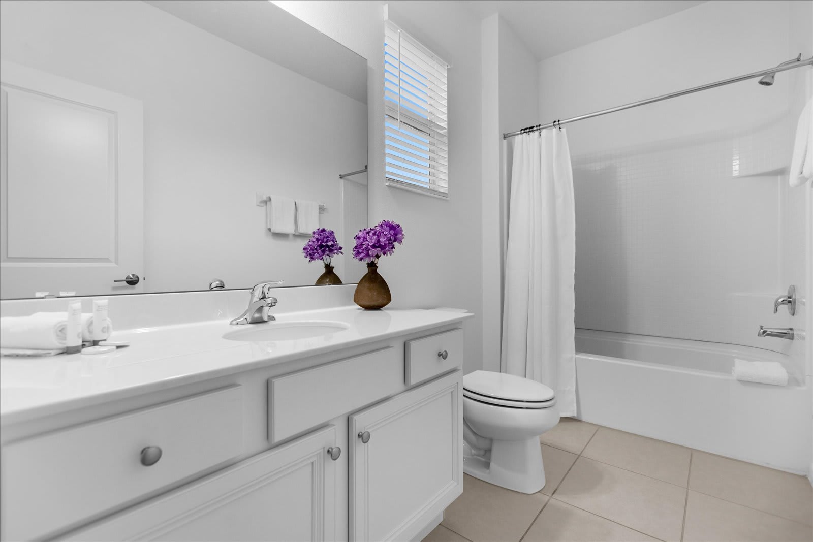 A bright, minimalist bathroom with white walls, a single vanity, and a combination shower/bathtub. The vanity features a white countertop, chrome faucet, and white shaker-style cabinets, with purple floral decor adding a pop of color.