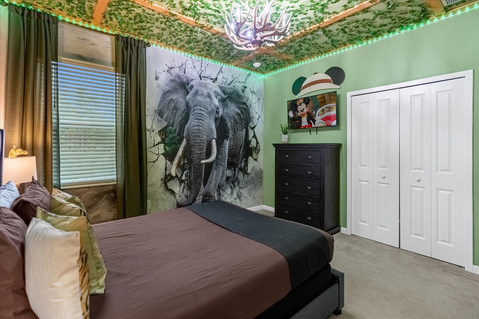 Themed bedroom with a large bed, featuring a dramatic wall mural of an elephant breaking through a wall, a jungle-themed ceiling with a chandelier, and a TV mounted above a black dresser.
