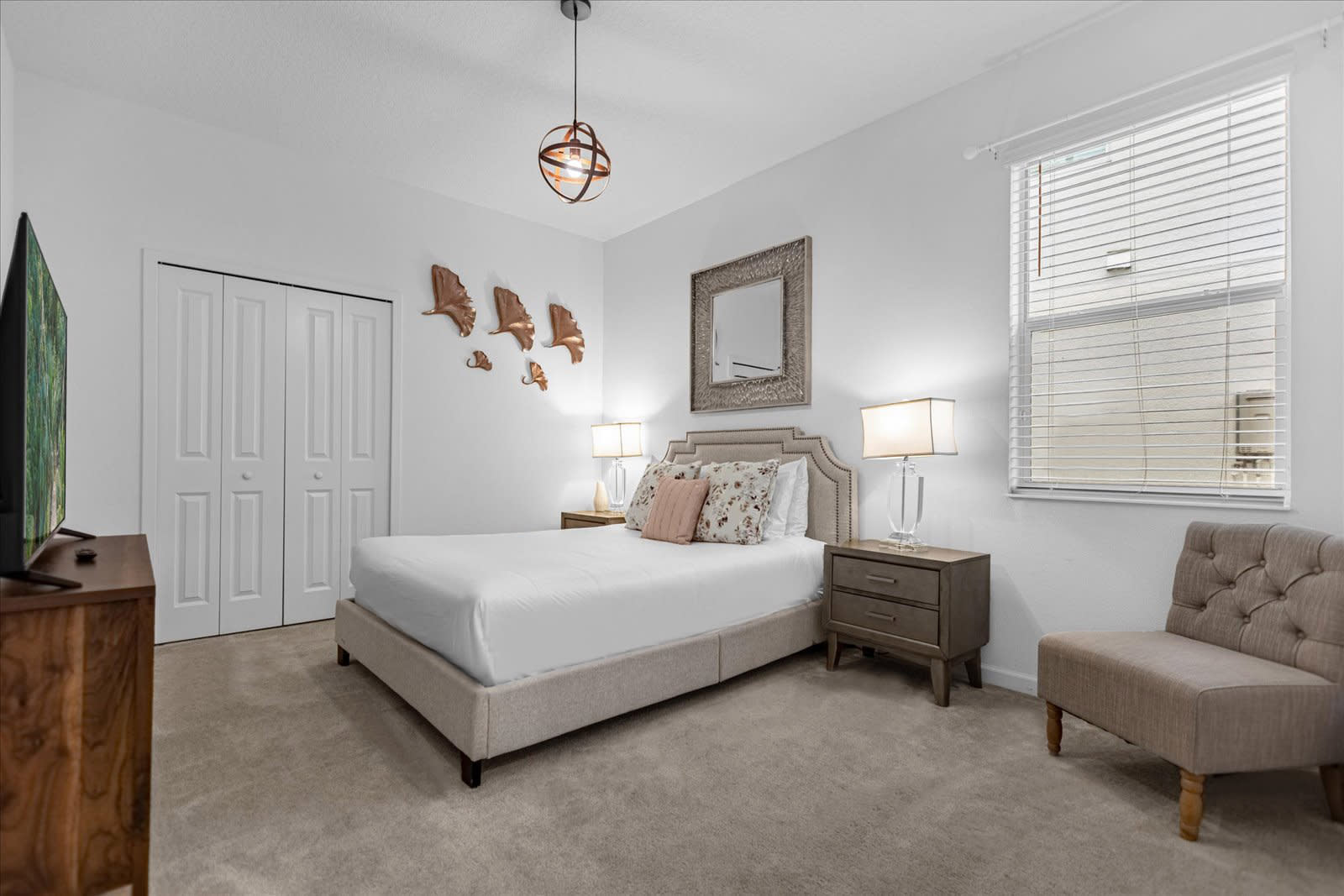 Cozy bedroom with a queen-sized bed, a tufted headboard, two nightstands with lamps, and a large TV on a wooden dresser. The room features soft carpeted flooring, a window with blinds, and decorative wall art.