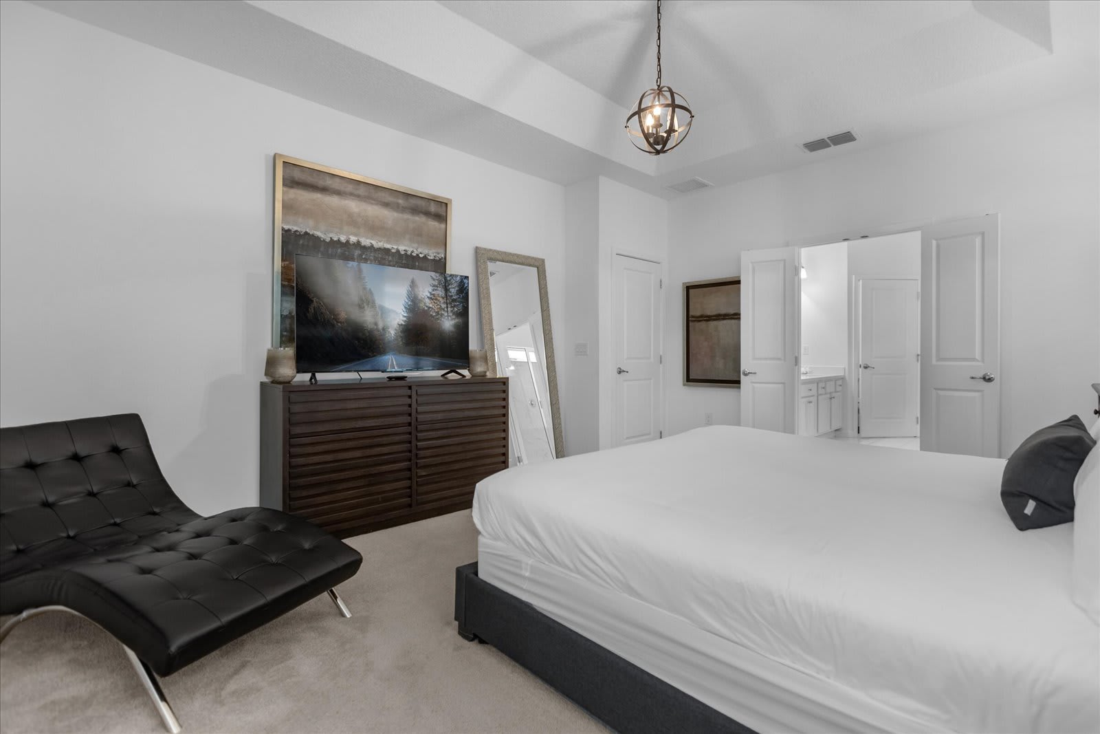 View of the master bedroom showing a large flat-screen TV on a dark wood dresser, a black chaise lounge, and a full-length mirror, with a glimpse of the en-suite bathroom through an open door.