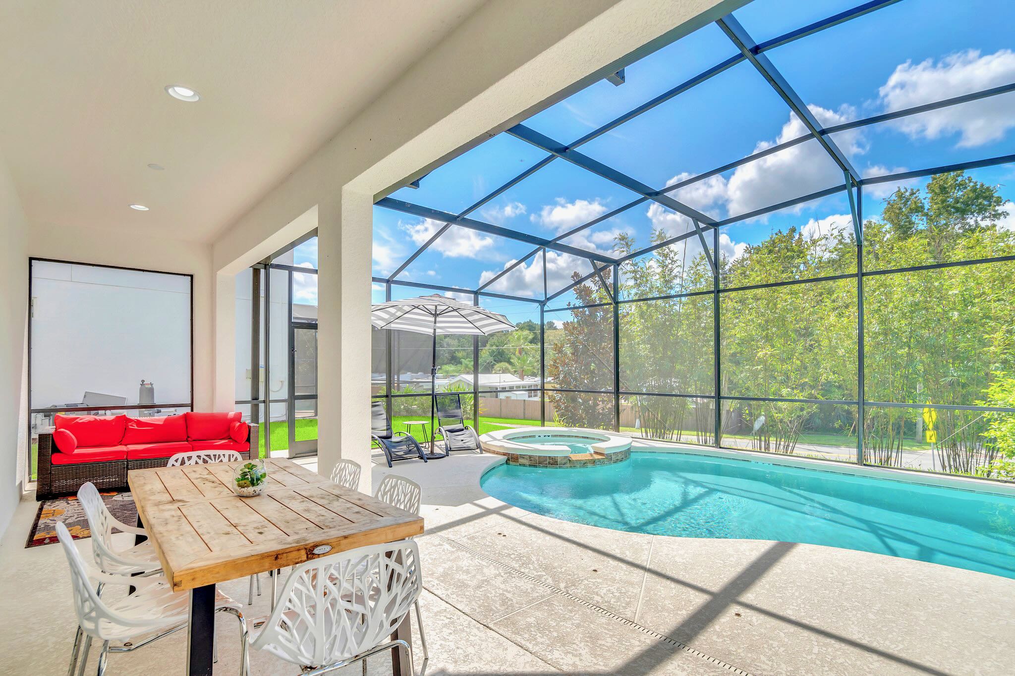 Covered outdoor patio area adjacent to a screened private pool, featuring a long wooden dining table with white chairs and a red outdoor sofa. Lush greenery is visible beyond the screen, creating a serene al fresco dining and lounging space.