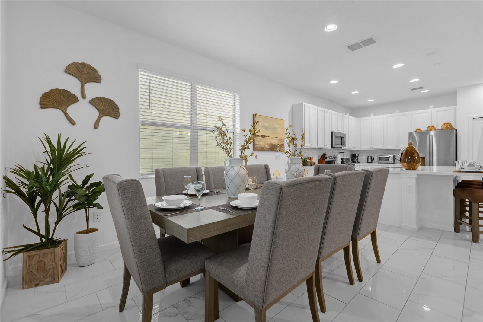 Bright dining area featuring a long rectangular wooden table with eight gray upholstered chairs, set on white tiled floors, with a large window and a modern white kitchen visible in the background.