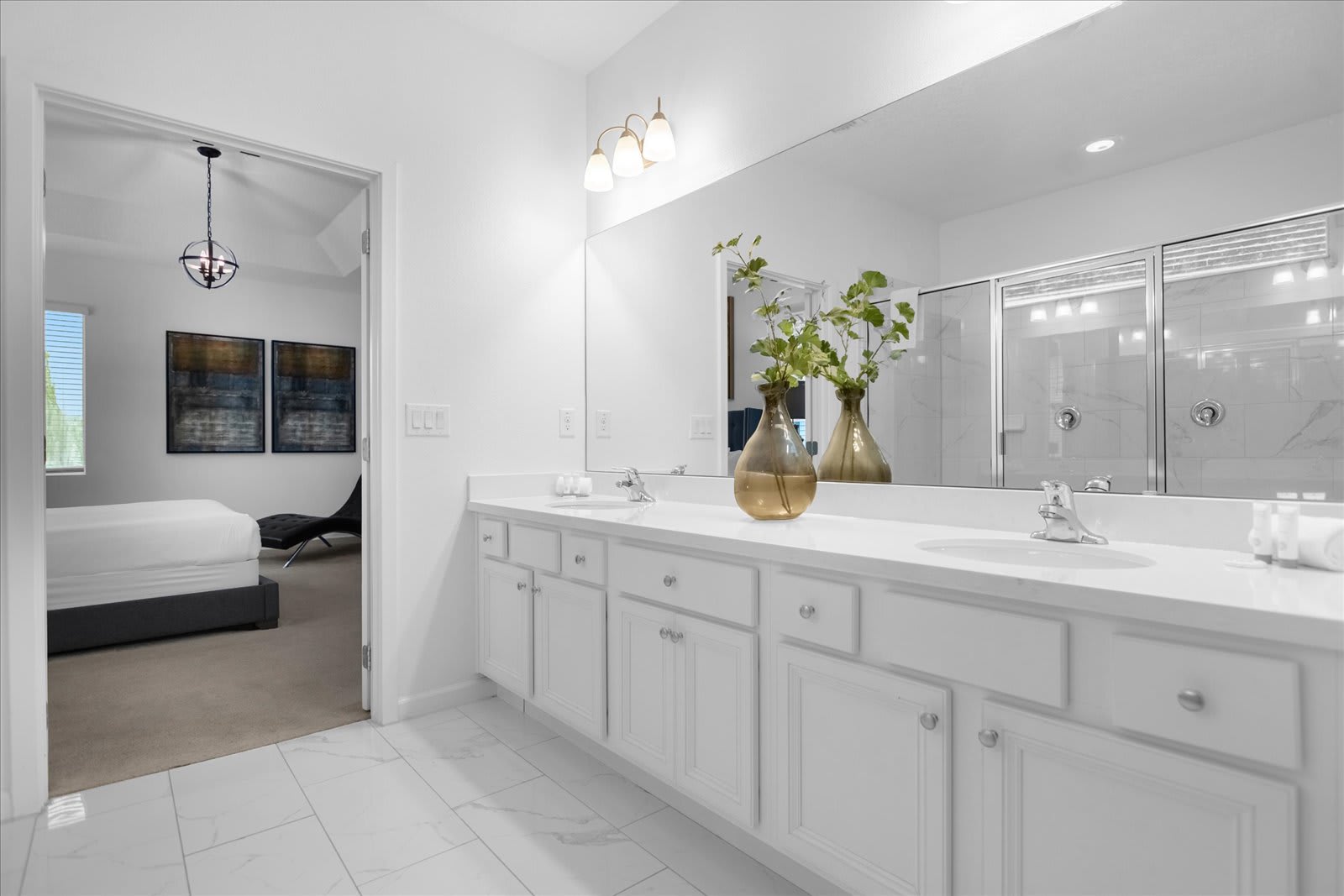 Spacious white master bathroom featuring a double vanity with white countertops, white cabinets, a large mirror, and a glass-enclosed shower. A bedroom is visible through an open doorway, suggesting an en-suite layout.