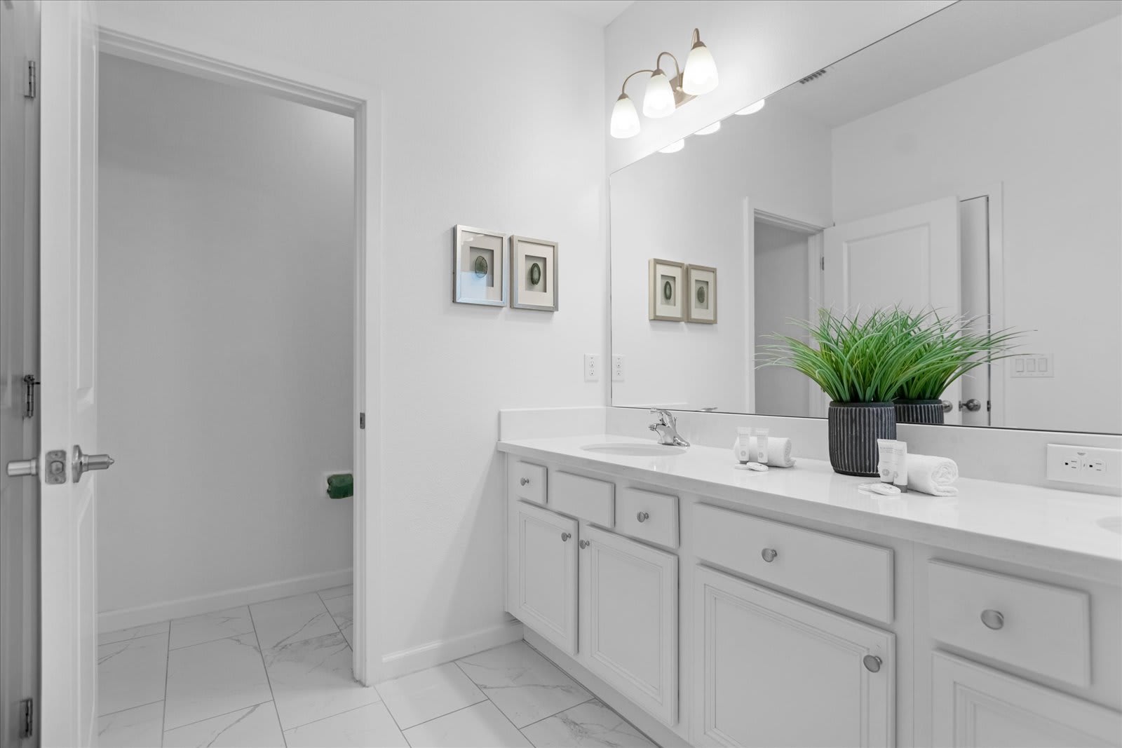 Clean white bathroom featuring a double vanity with white countertops, a large mirror, and white cabinets. A separate toilet room is visible through an open doorway on the left, complementing the bright, minimalist design.