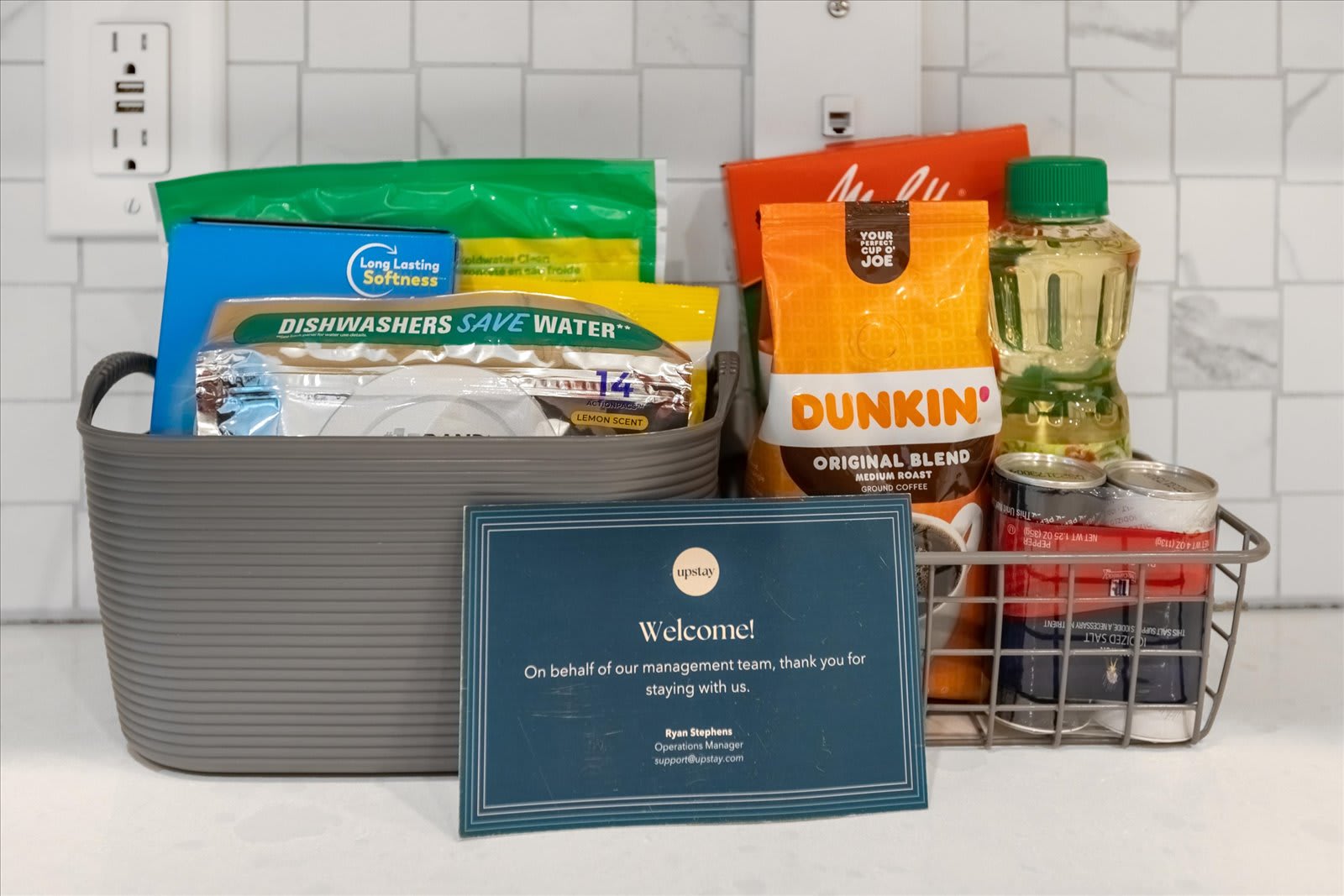 A thoughtful welcome basket on a kitchen counter, containing various household essentials like dish soap, coffee, and cooking oil, alongside a "Welcome!" card from the property management.
