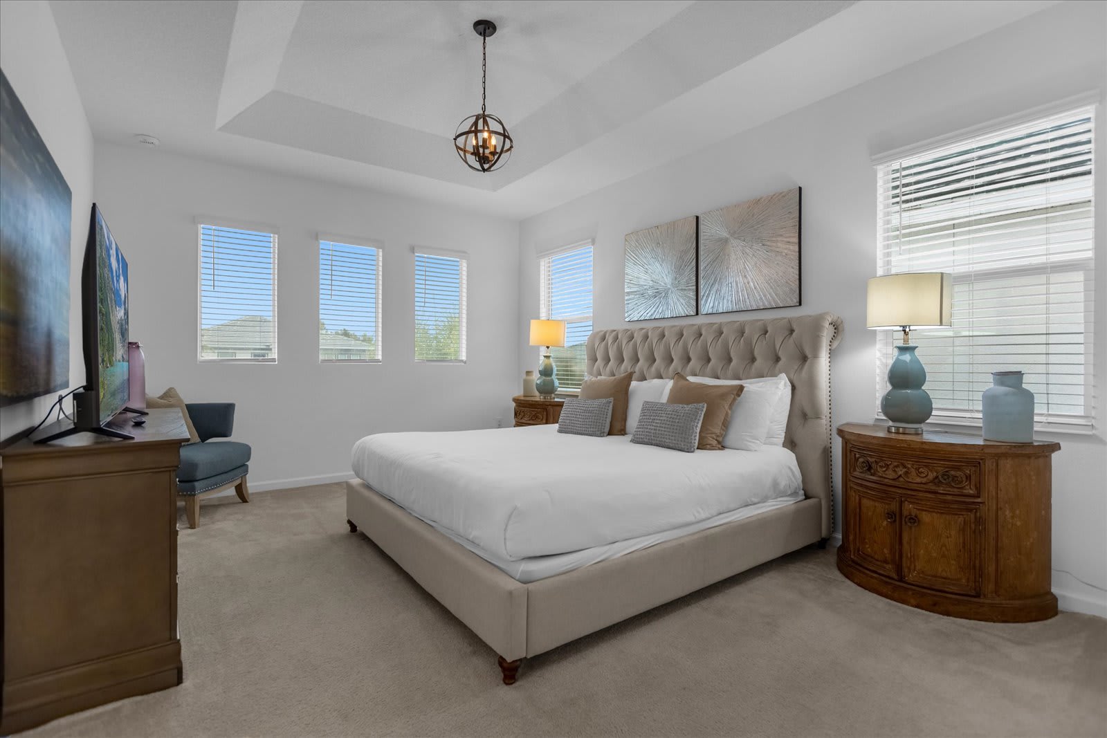 Spacious master bedroom featuring a king-sized bed with a tufted headboard, two wooden nightstands with lamps, and multiple windows with blinds. The room has soft carpeted flooring and a tray ceiling with a decorative chandelier.