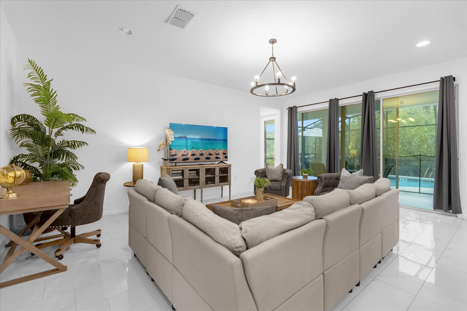Modern living room featuring a large L-shaped sectional sofa, a flat-screen TV on a wooden console, and sliding glass doors leading to an outdoor pool area, with light tile flooring throughout.