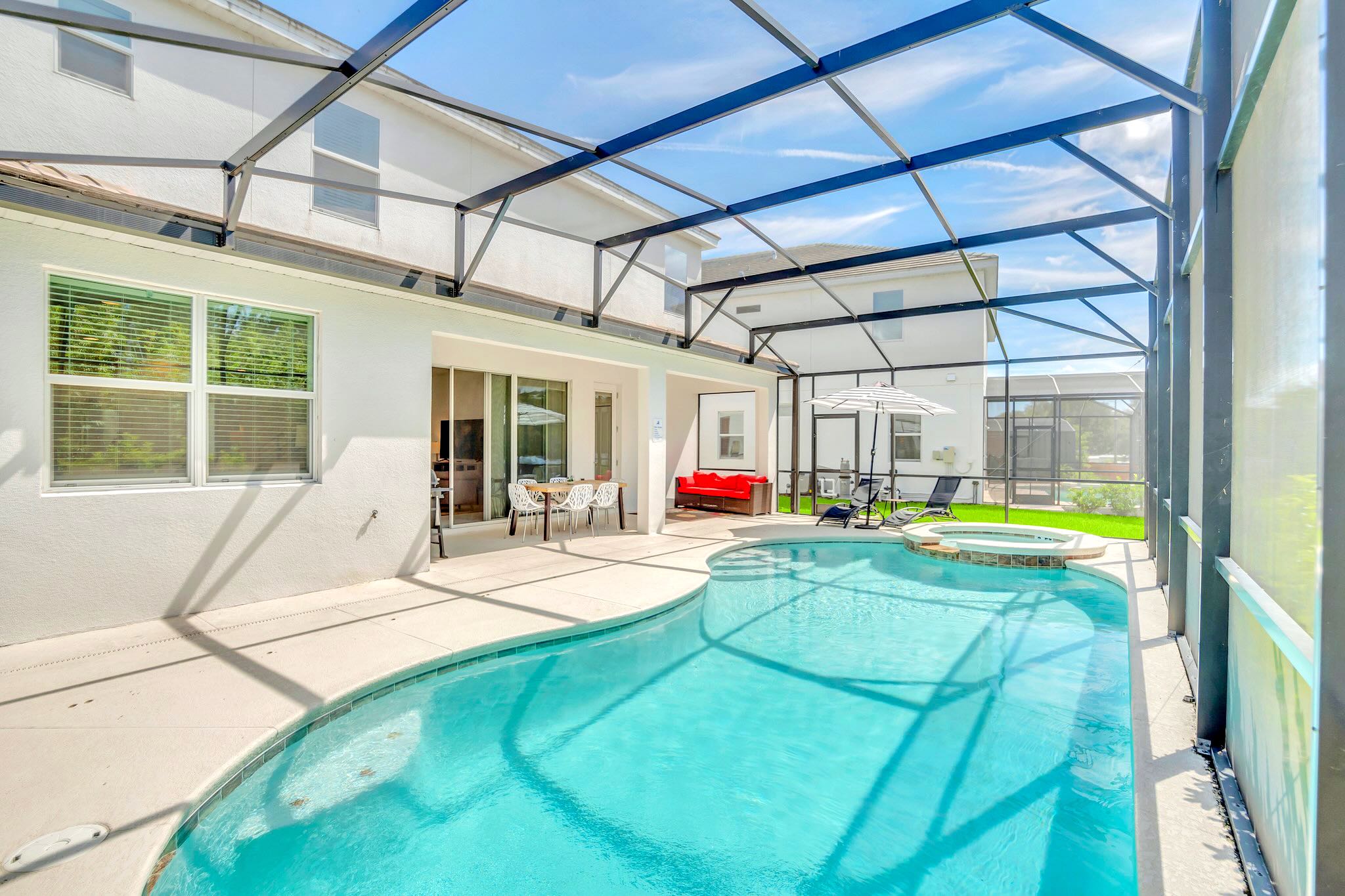 Screened-in private pool and hot tub area with lounge chairs, a striped umbrella, and an outdoor seating area with a red sofa. The white house exterior and large windows are visible, offering a bright and relaxing space.