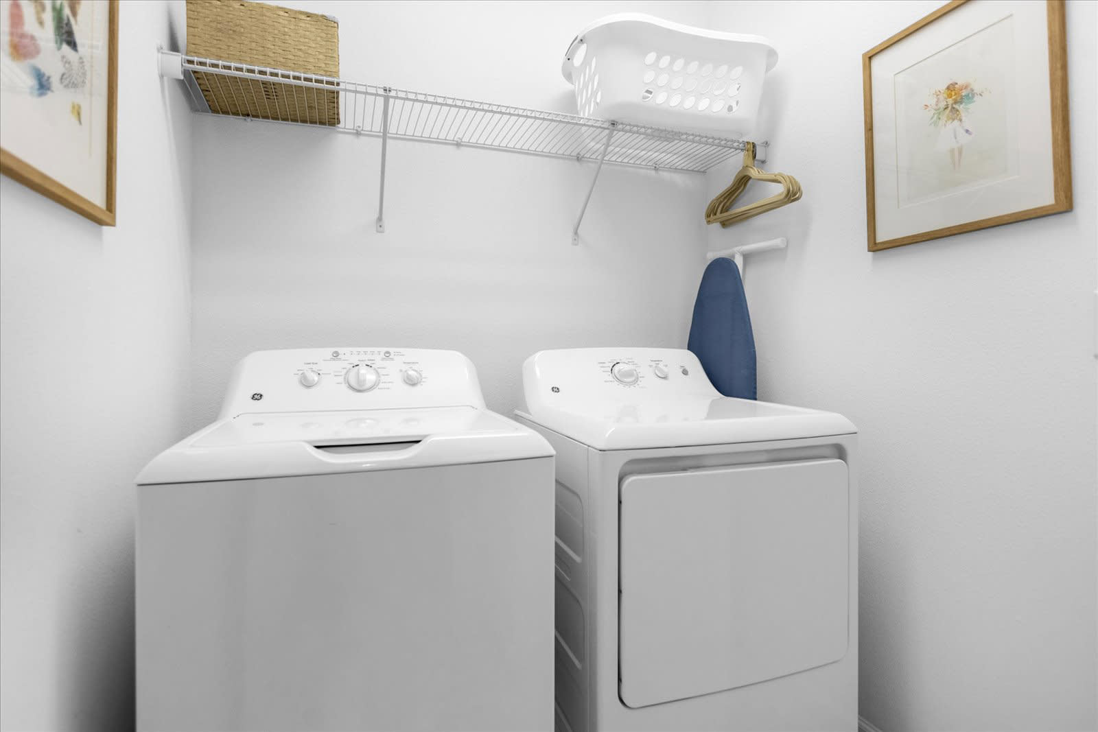 A compact laundry room with white walls, featuring a white top-load washing machine and a front-load dryer. A wire shelf above provides storage, with a laundry basket and ironing board visible.