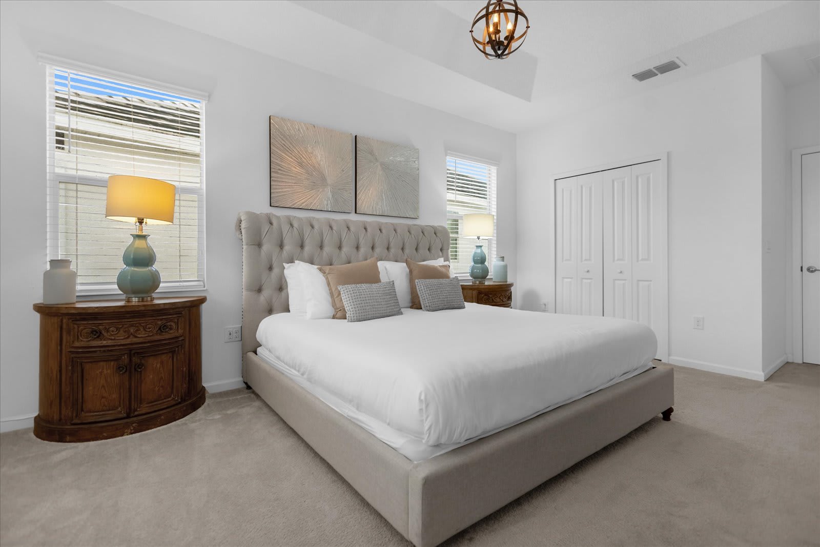 Elegant master bedroom with a king-sized bed, a plush tufted headboard, two ornate wooden nightstands with lamps, and a large closet with white bi-fold doors. The room is carpeted and features a tray ceiling with a chandelier.