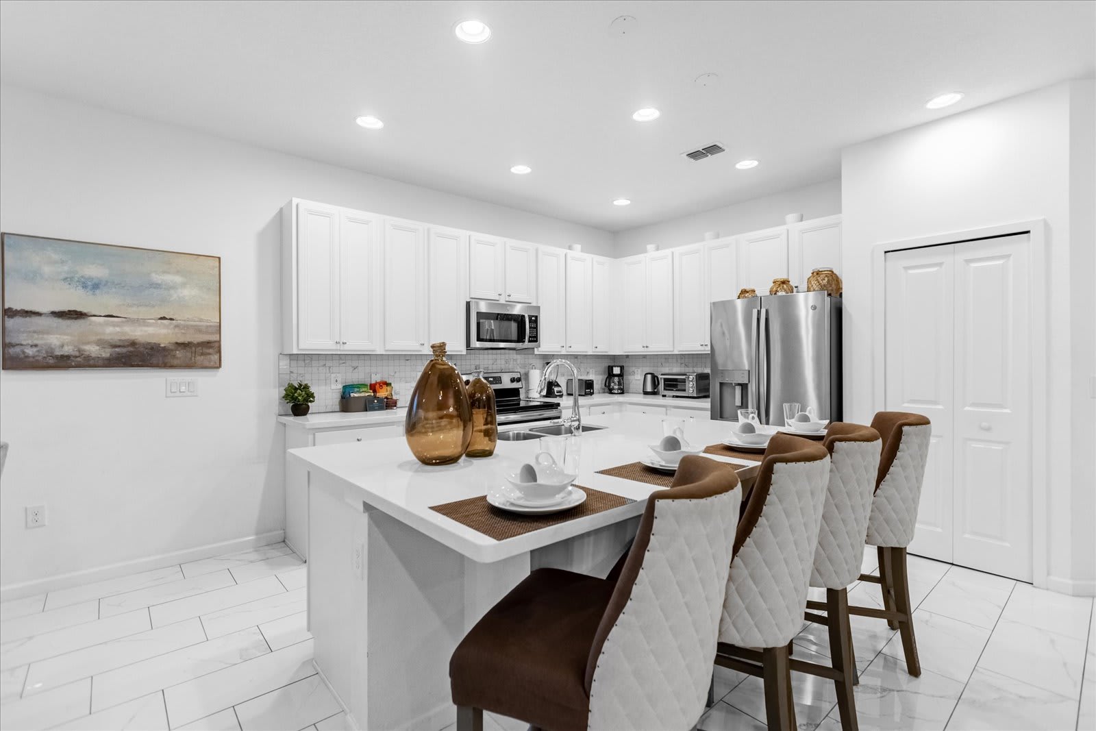 Contemporary kitchen with a large white quartz island and bar stools, white shaker cabinets, stainless steel appliances including a refrigerator and microwave, and a white subway tile backsplash.