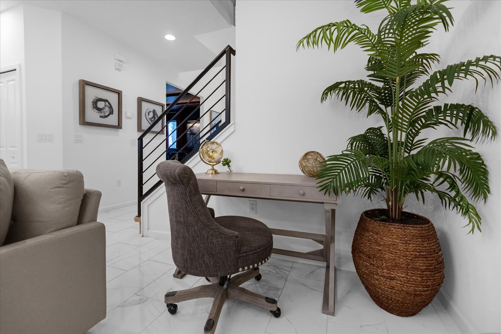 Cozy office nook with a light wooden desk and a brown upholstered swivel chair, positioned next to a modern staircase with black railings, and a large potted palm plant in a woven basket.