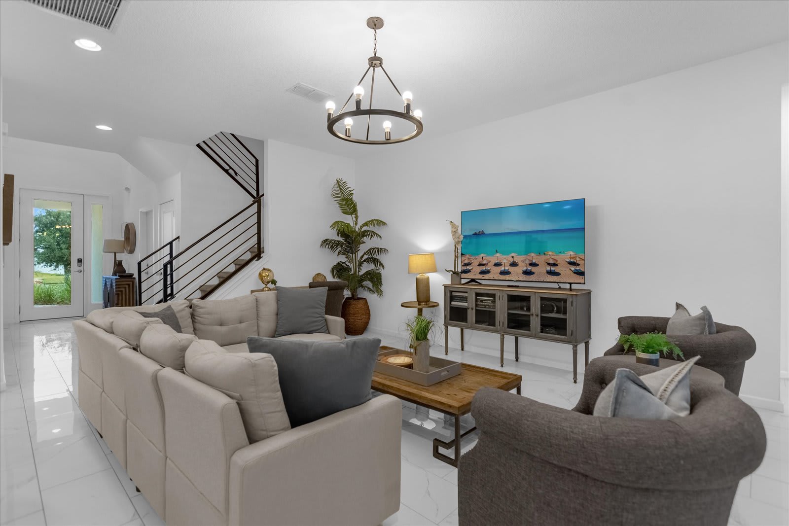 Bright and spacious living room with white walls, light tile flooring, a large beige sectional sofa, and two gray armchairs. A rustic wooden coffee table, modern chandelier, and large flat-screen TV create a comfortable gathering space.
