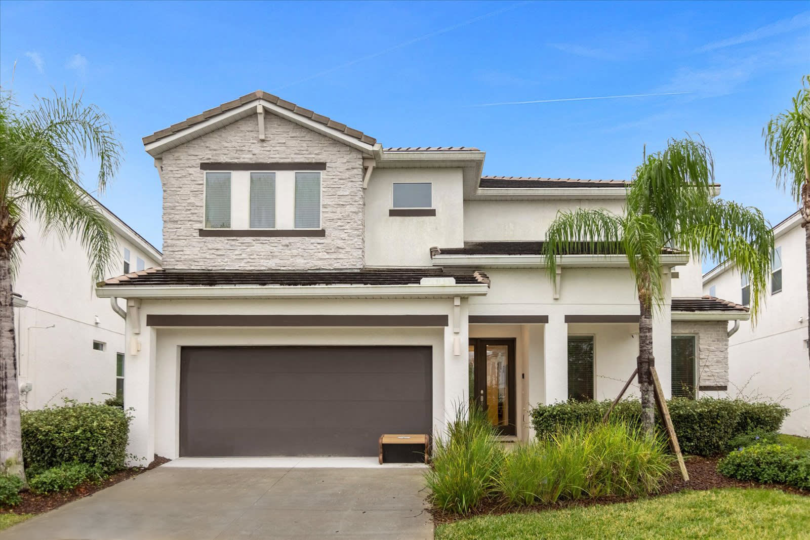Modern two-story house with a light-colored stone and stucco facade, a dark garage door, and a paved driveway. Lush green landscaping with palm trees frames the entrance under a clear blue sky.