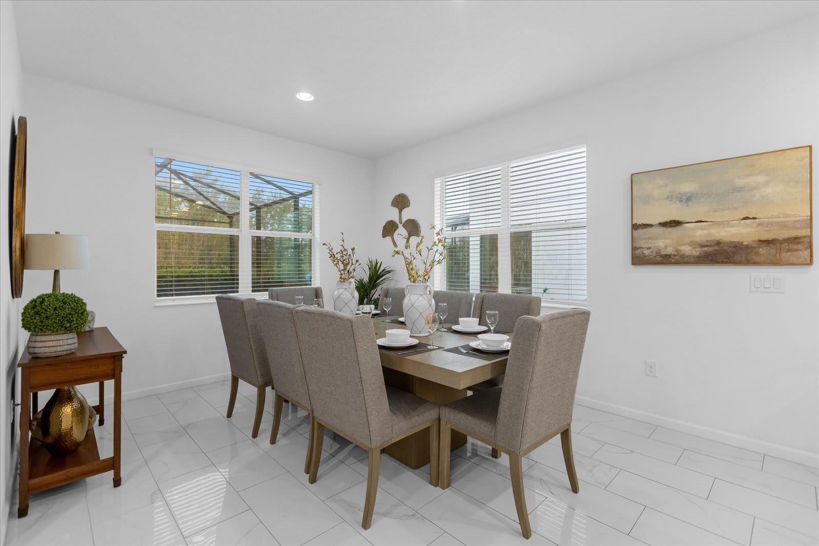 Spacious dining room with a light wood table and eight gray upholstered chairs on white tiled floors, illuminated by two large windows overlooking a screened outdoor area, complemented by abstract wall art.