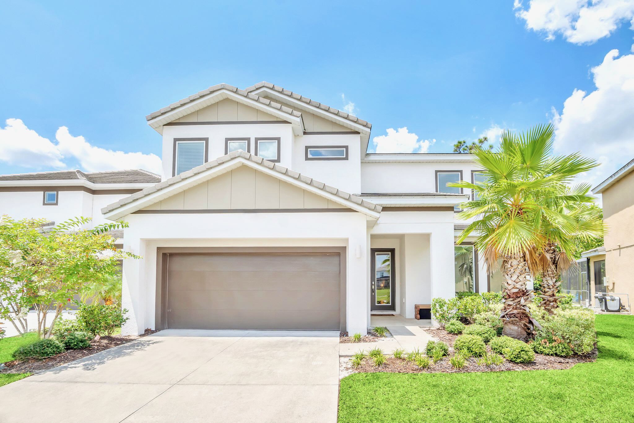 The modern two-story house features a white and gray facade with a brown garage door, a paved driveway, and a well-maintained green lawn with palm trees and landscaping. The front door is visible through a small porch.