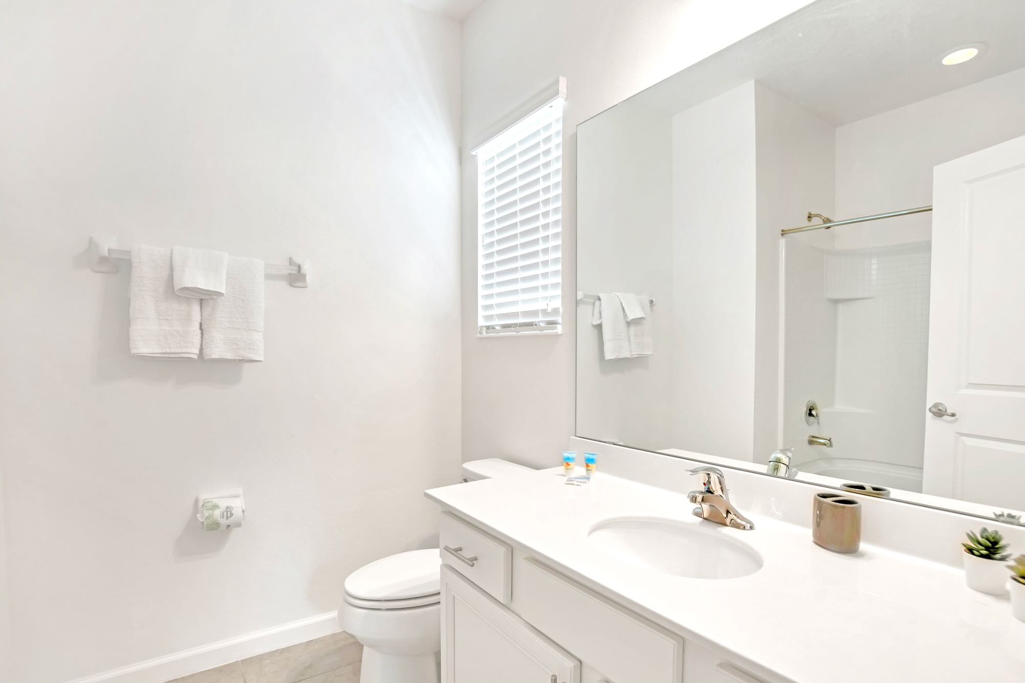 Clean bathroom with a white single vanity, a toilet, and a bathtub with a light-colored shower curtain, featuring a large mirror and light gray floor tiles.