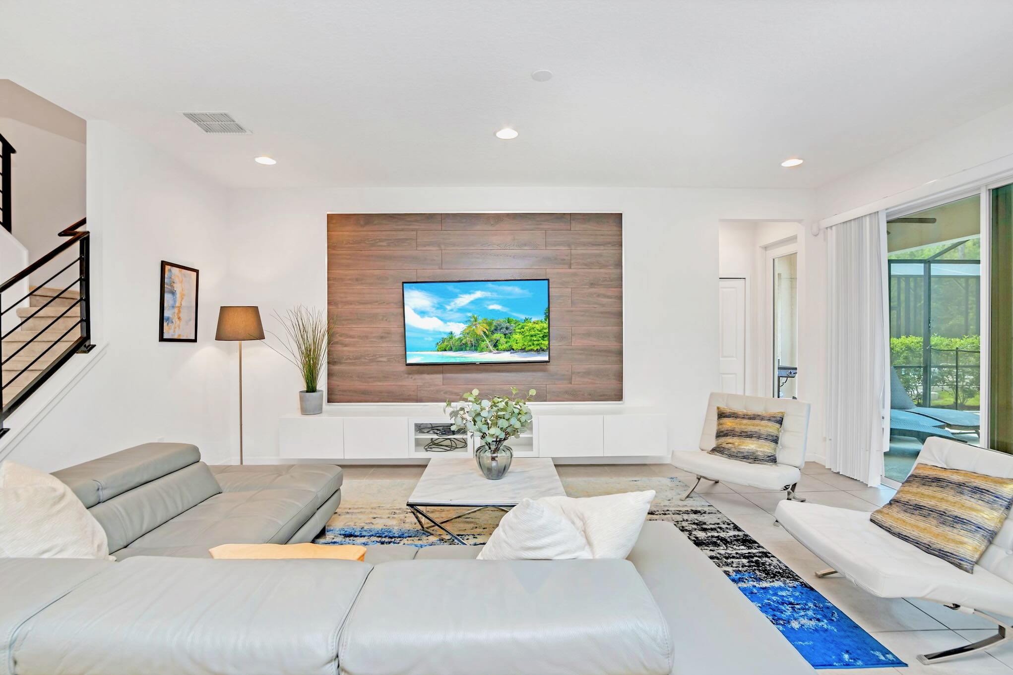 A modern living room features a large wall-mounted TV set against a wood-paneled accent wall, a gray sectional sofa, and two white armchairs. A blue and gold abstract rug adds a pop of color to the tiled floor.