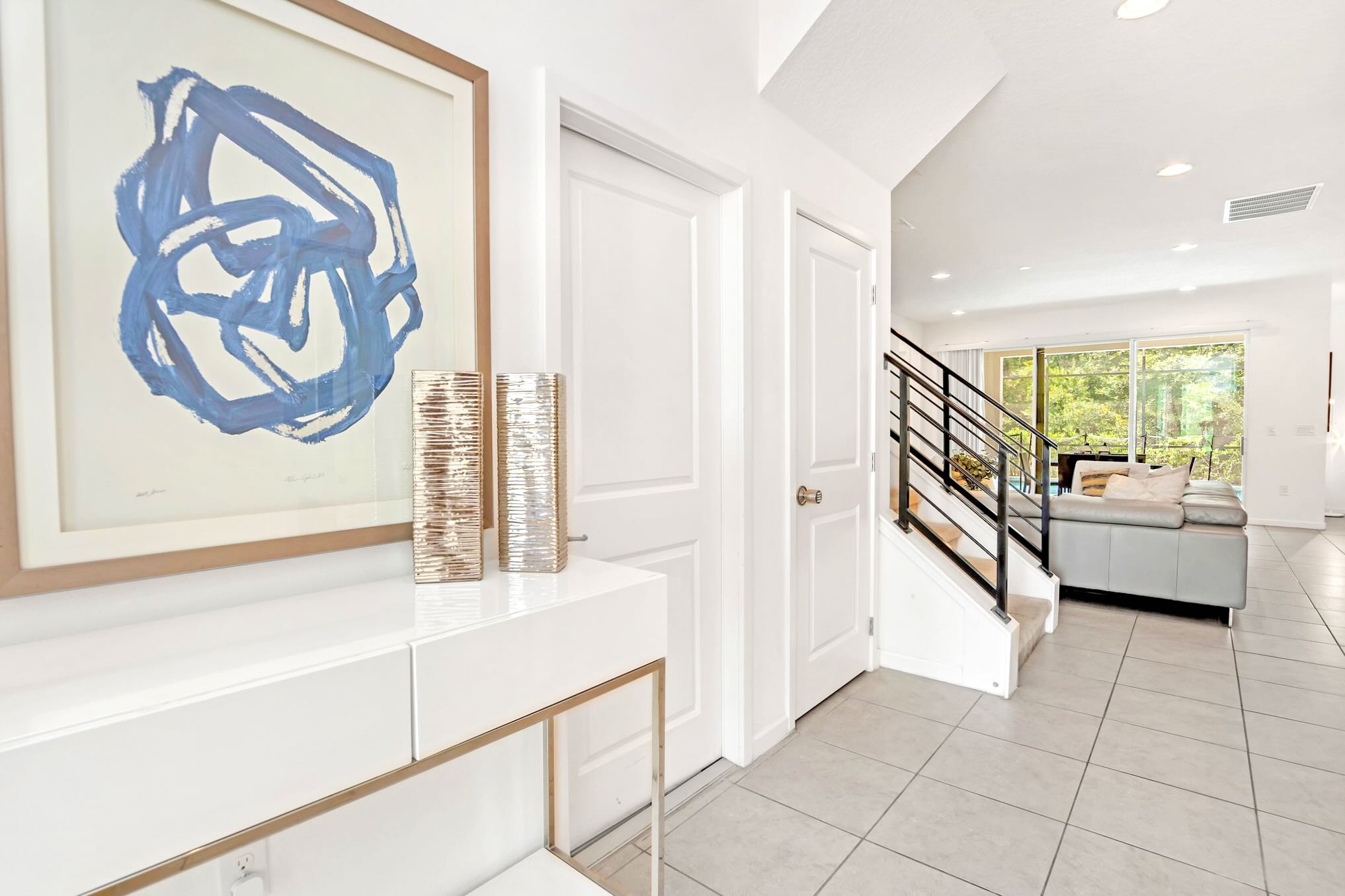 Bright foyer with a white console table, decorative vases, and a large framed abstract blue artwork. White doors and a modern staircase with black railings lead to the upper floor, with tile flooring throughout.
