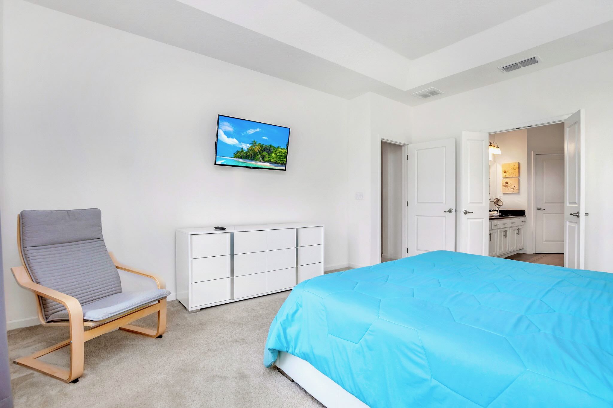 View of bedroom-1 showing a wall-mounted TV above a white dresser, and a modern gray lounge chair. The light gray carpet and white walls continue from the previous view, with a doorway leading to an en-suite bathroom.