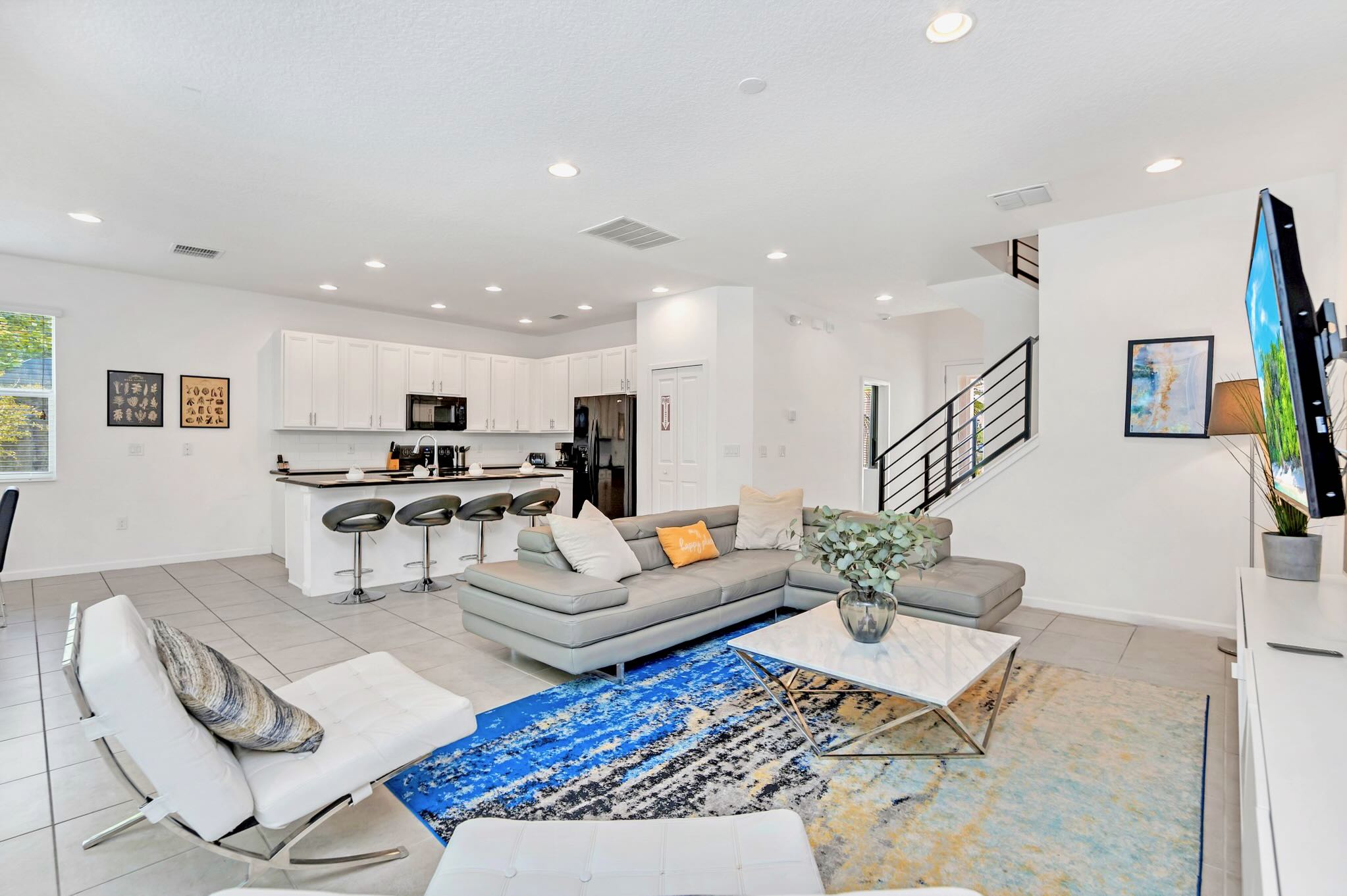 An open-concept living area features a large gray sectional sofa, a white marble coffee table, and a vibrant blue and gold abstract rug, with a modern white kitchen with black countertops visible in the background. A staircase with black metal railings is also visible.